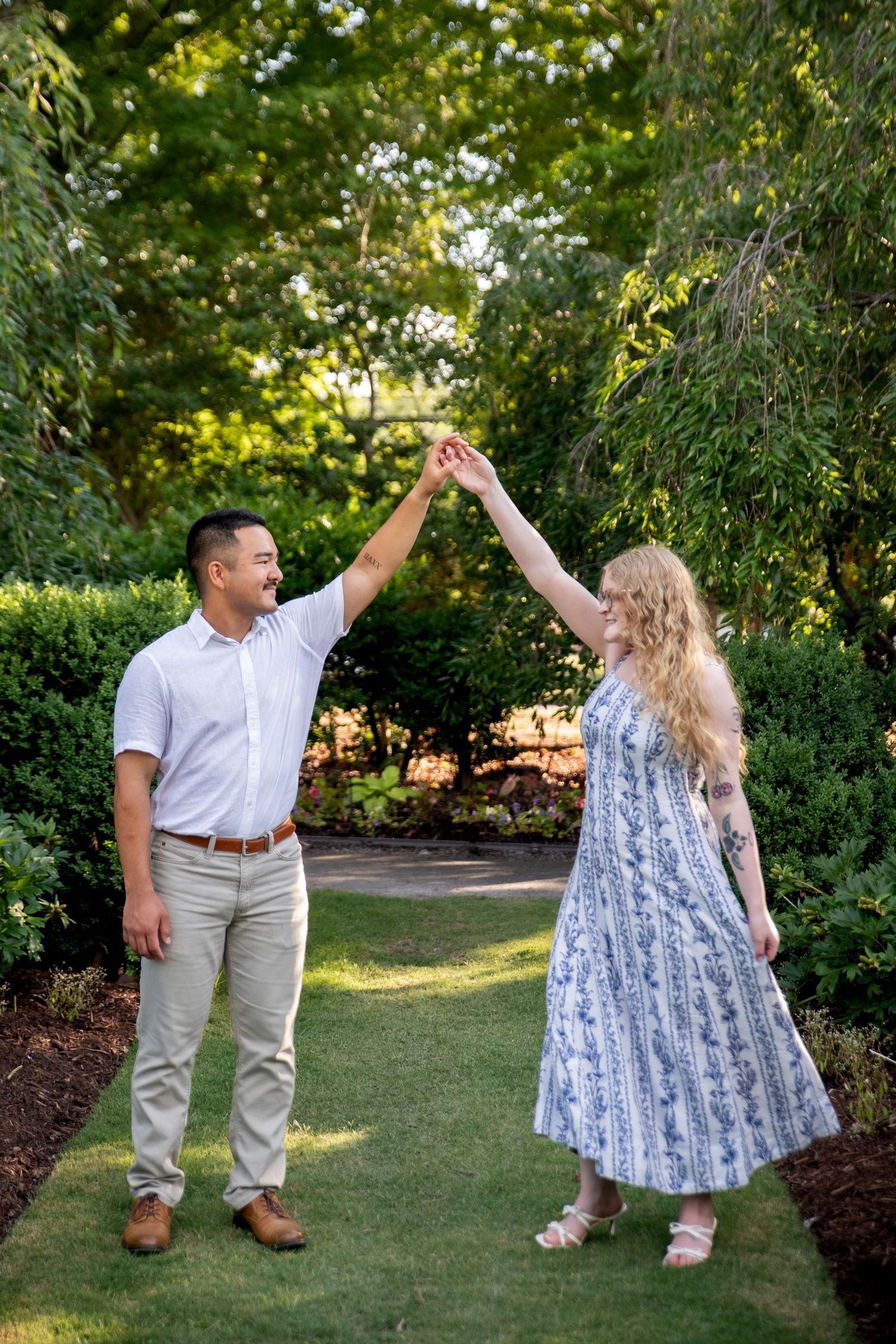 A couple dances outdoors in a lush garden, man in white shirt and woman in blue floral dress, holding hands raised.
