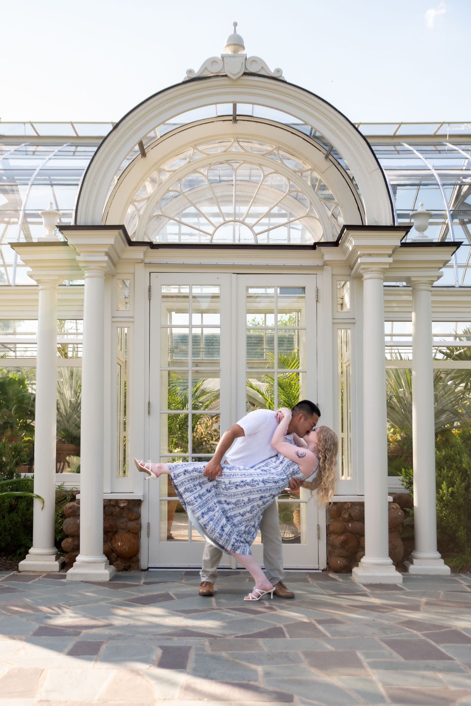 Couple sharing a romantic dip kiss in front of an elegant white greenhouse conservatory with arched windows.