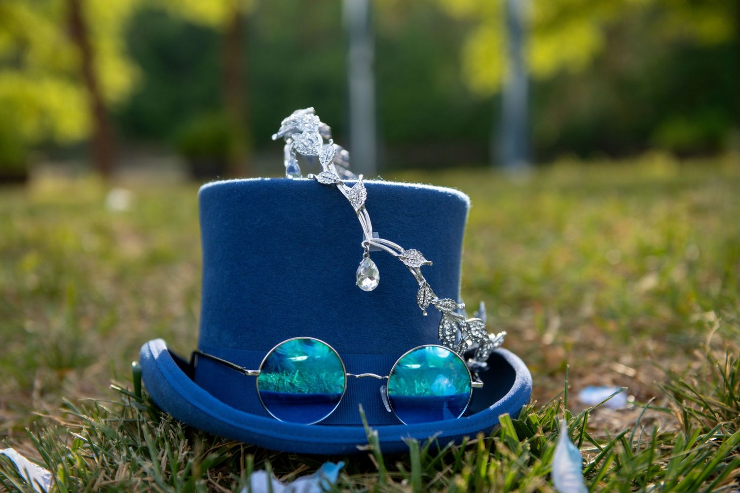 A blue top hat with round teal sunglasses and crystal ornament resting on green grass outdoors.