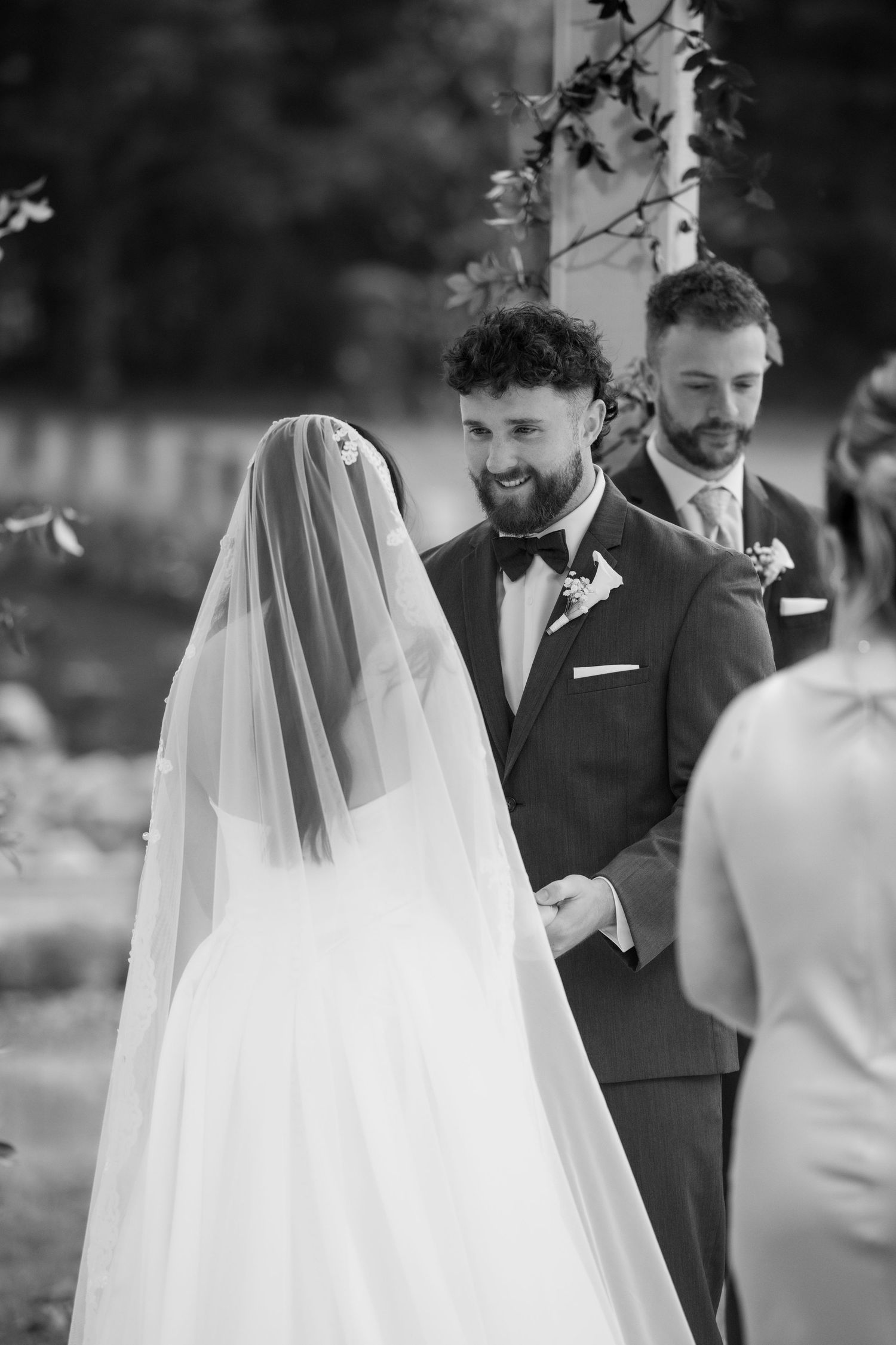 Black and white photo of a smiling groom holding hands with his bride in a veil during an outdoor wedding ceremony.