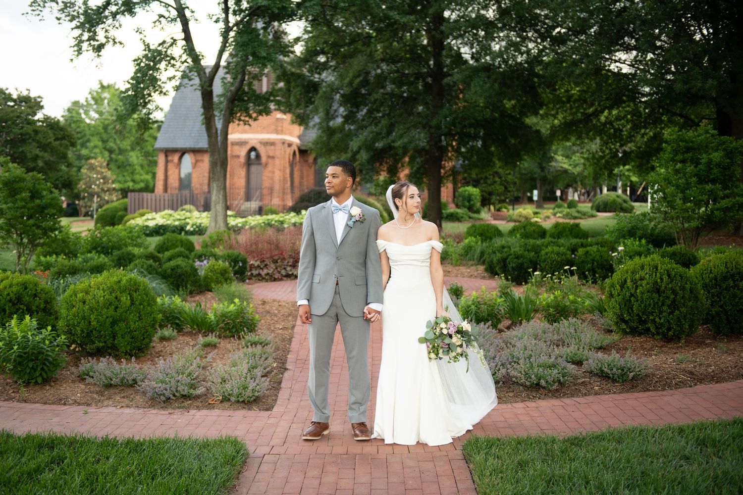 Bride and groom stand on brick path in lush garden with historic brick chapel backdrop on their wedding day.
