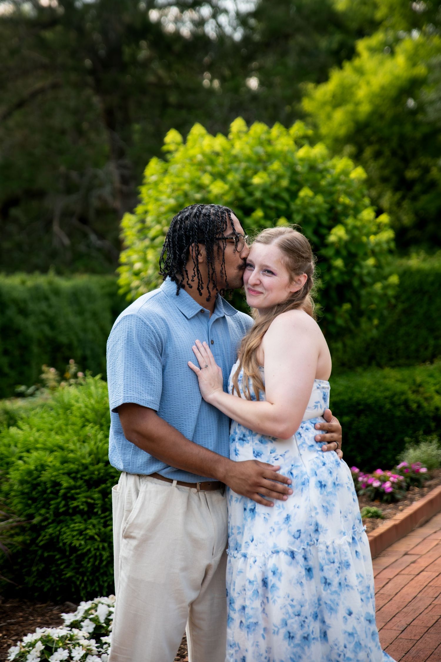 A couple shares a tender kiss in a lush garden, woman in floral dress smiling as man embraces her.