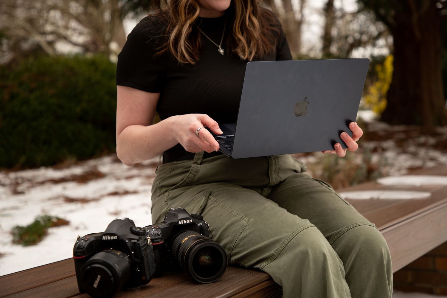 A person sits outdoors on a bench using a laptop while holding a camera in winter weather.
