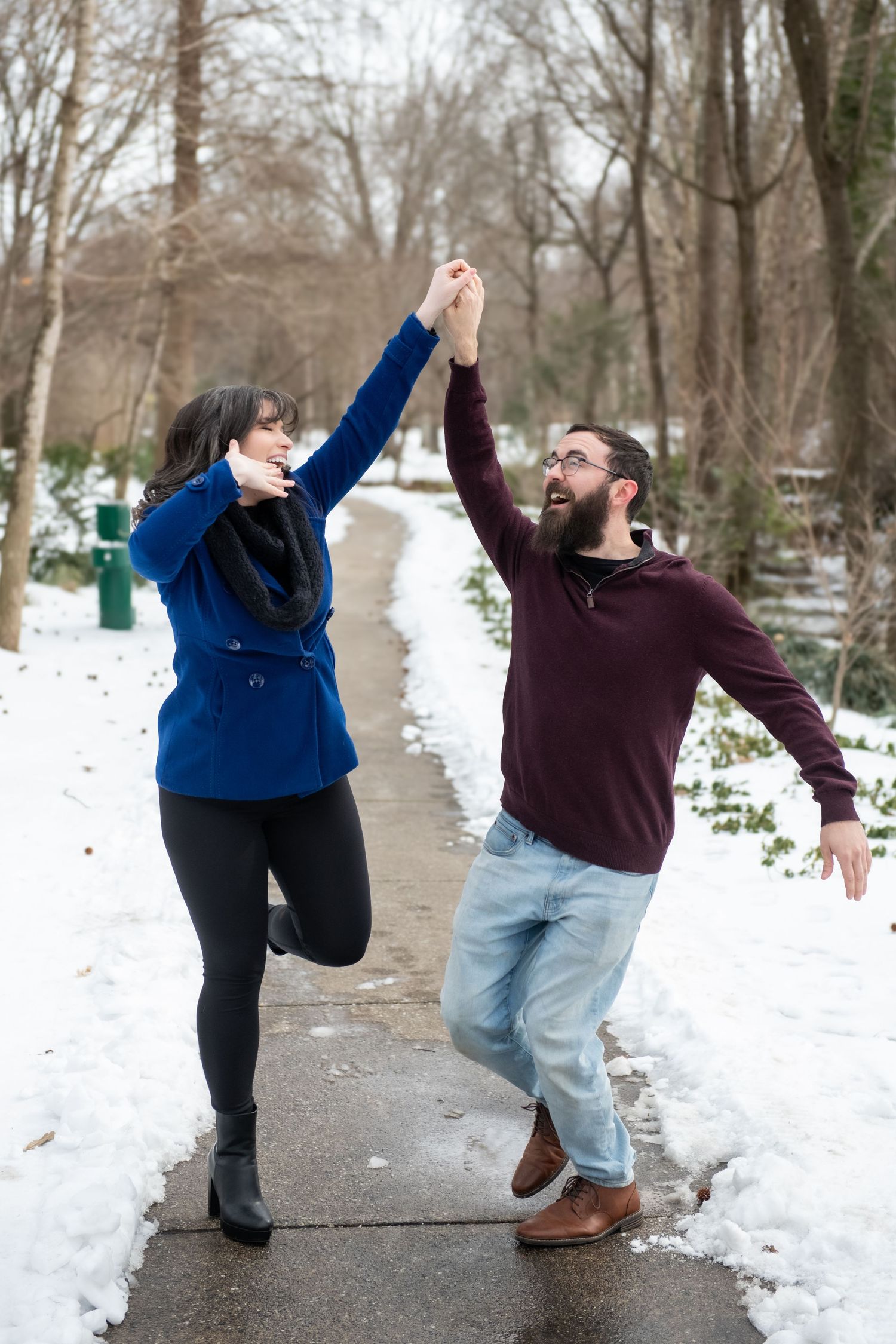 A couple strikes fun dancing poses on a snowy path during their winter photoshoot.