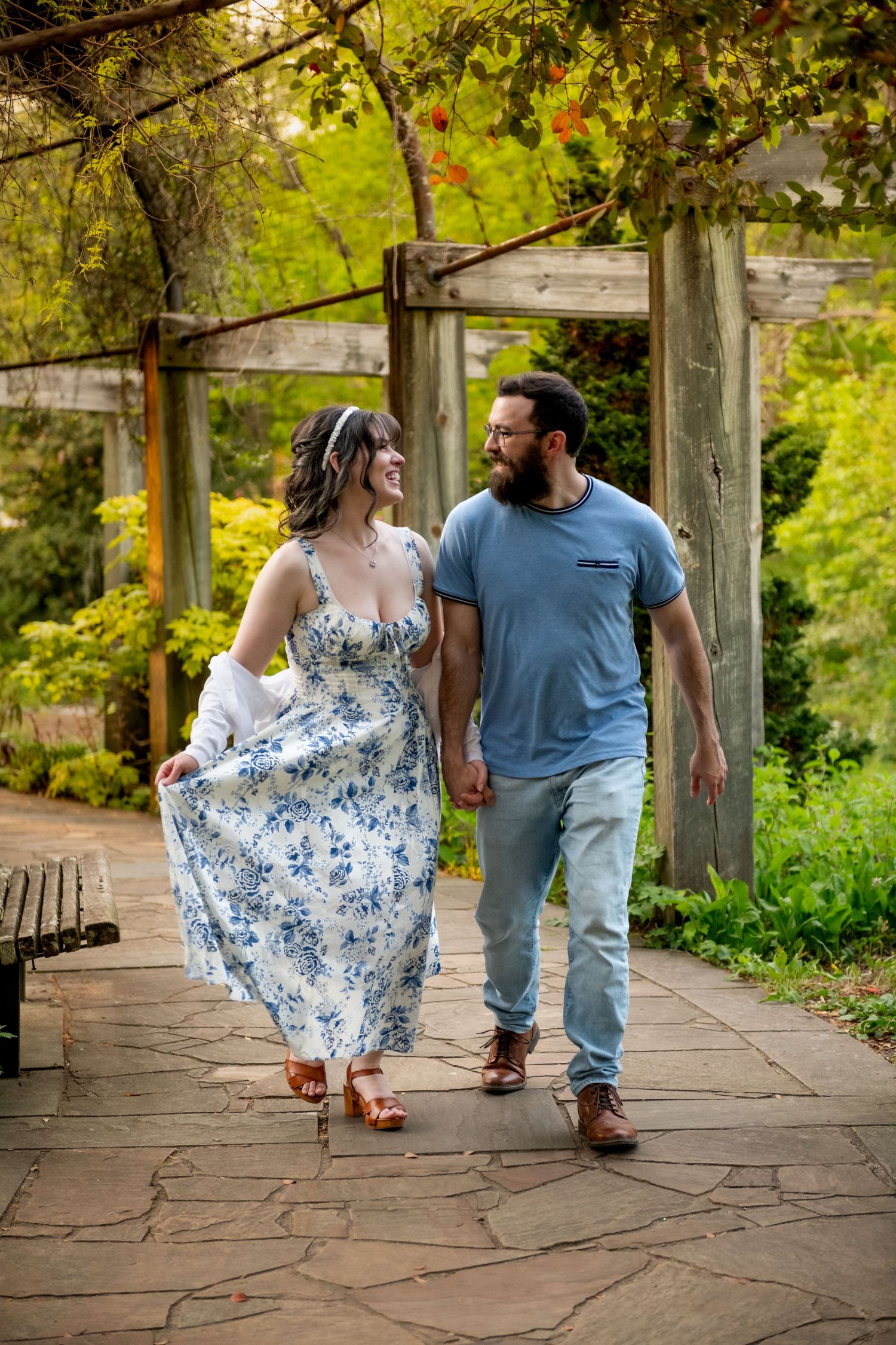 A smiling couple walks hand-in-hand through a lush garden pergola, woman in floral dress, man in blue t-shirt and jeans.