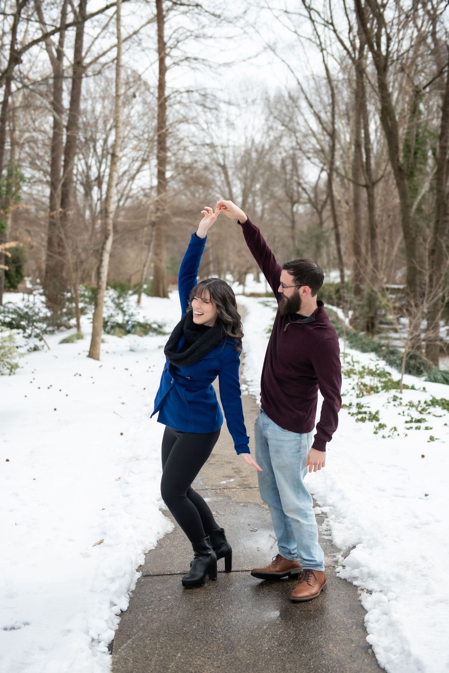 Two people dance playfully together in a snowy winter park setting with bare trees in the background.