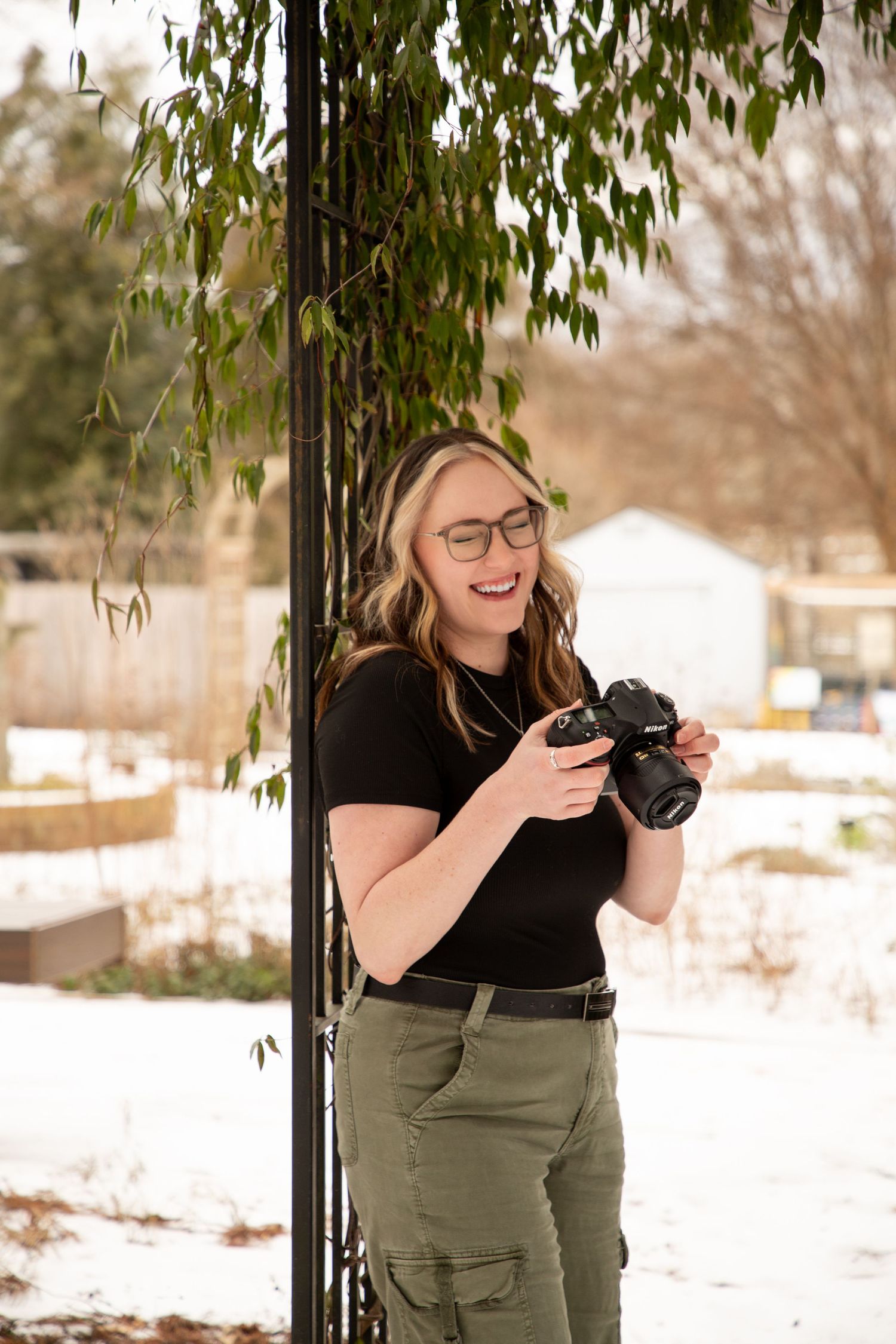 A photographer in a black shirt and olive pants laughs while holding a camera in a snowy outdoor setting.
