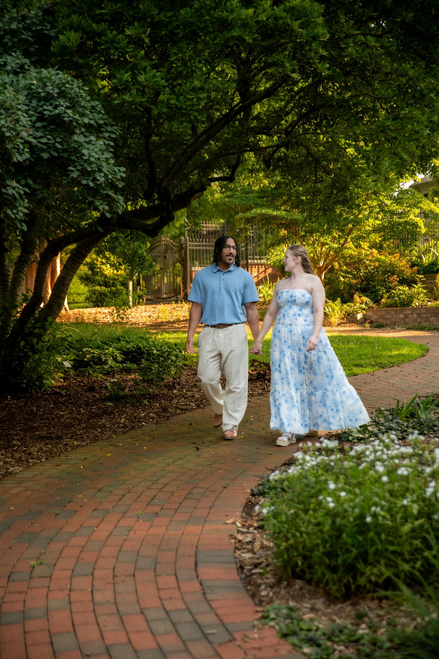 A couple holds hands walking on a brick path through a lush green garden, woman in floral dress, man in blue shirt.