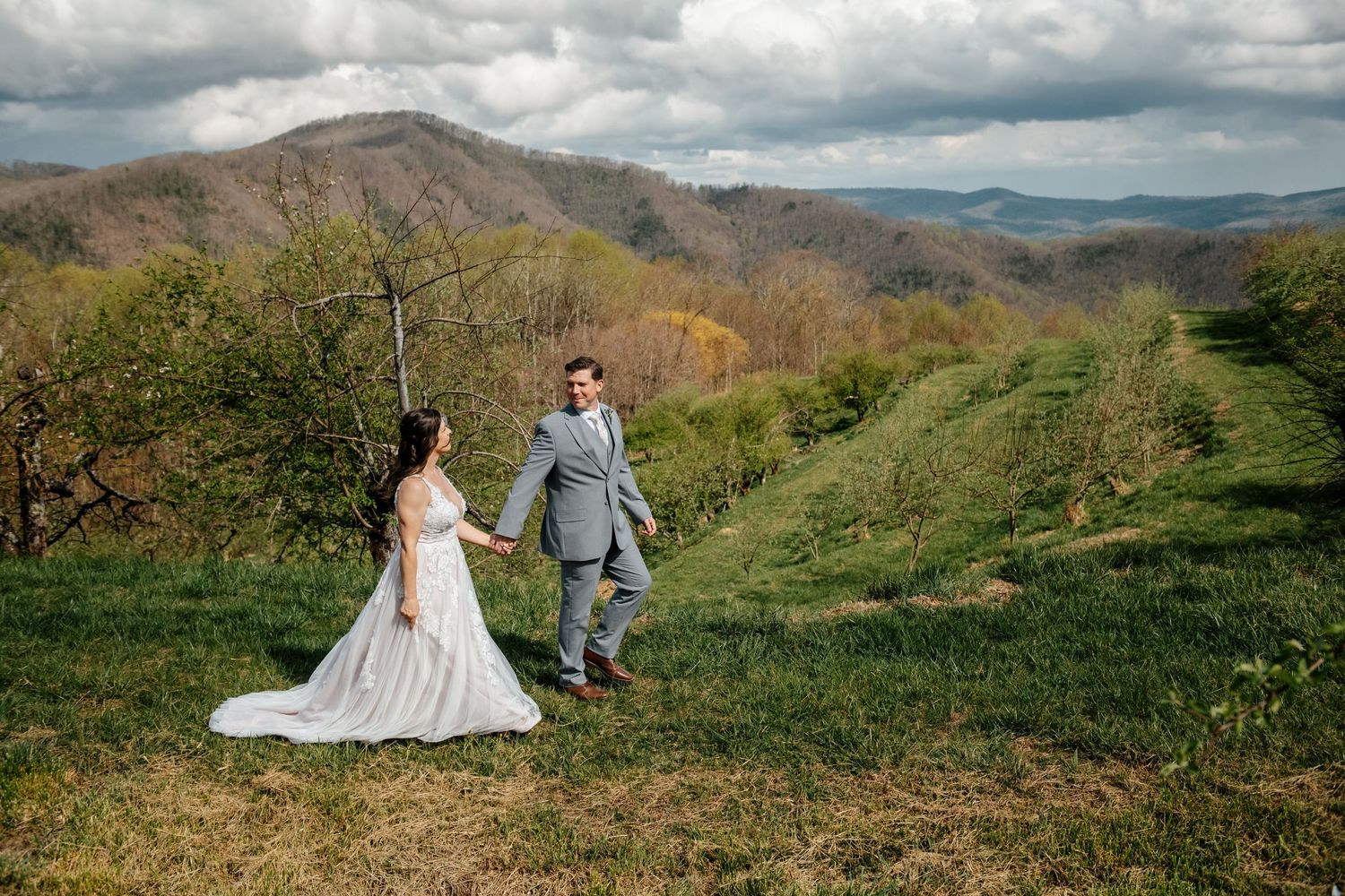 Bride and groom walk hand in hand through a lush green orchard with stunning mountain views.