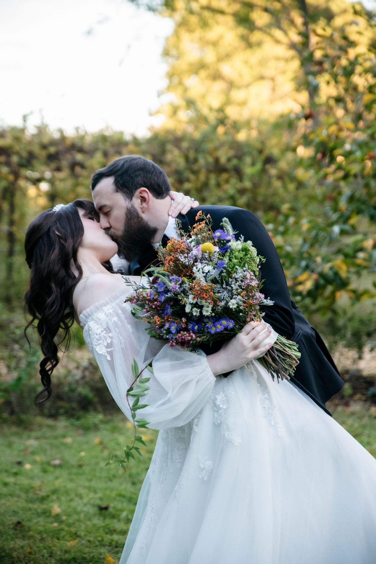 Bride in a white lace gown and groom in a dark suit share a dipped kiss outdoors, holding a wildflower bouquet in a sunlit garden.