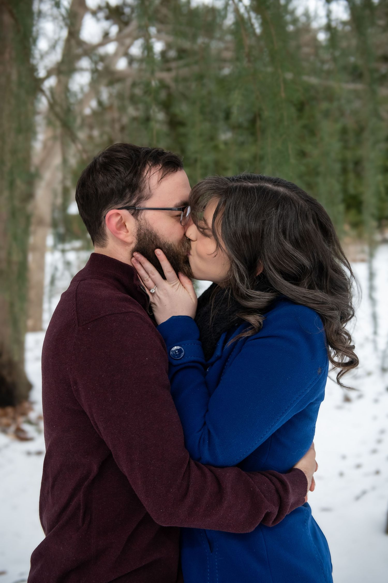 A romantic winter moment captured as a couple shares a tender kiss surrounded by snow-covered trees.