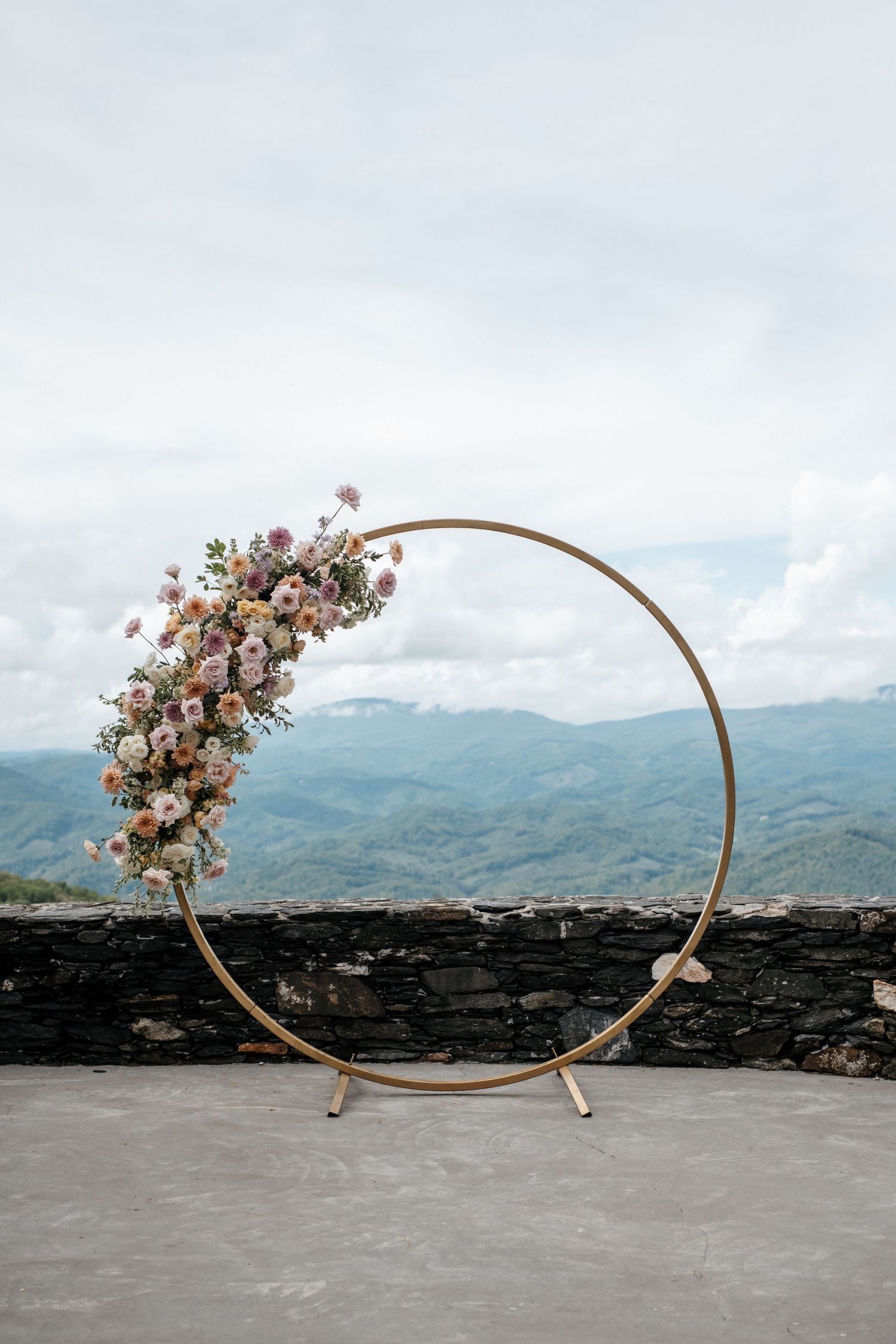 Gold circular wedding arch adorned with pink and white flowers, set on a stone terrace with scenic mountain views.