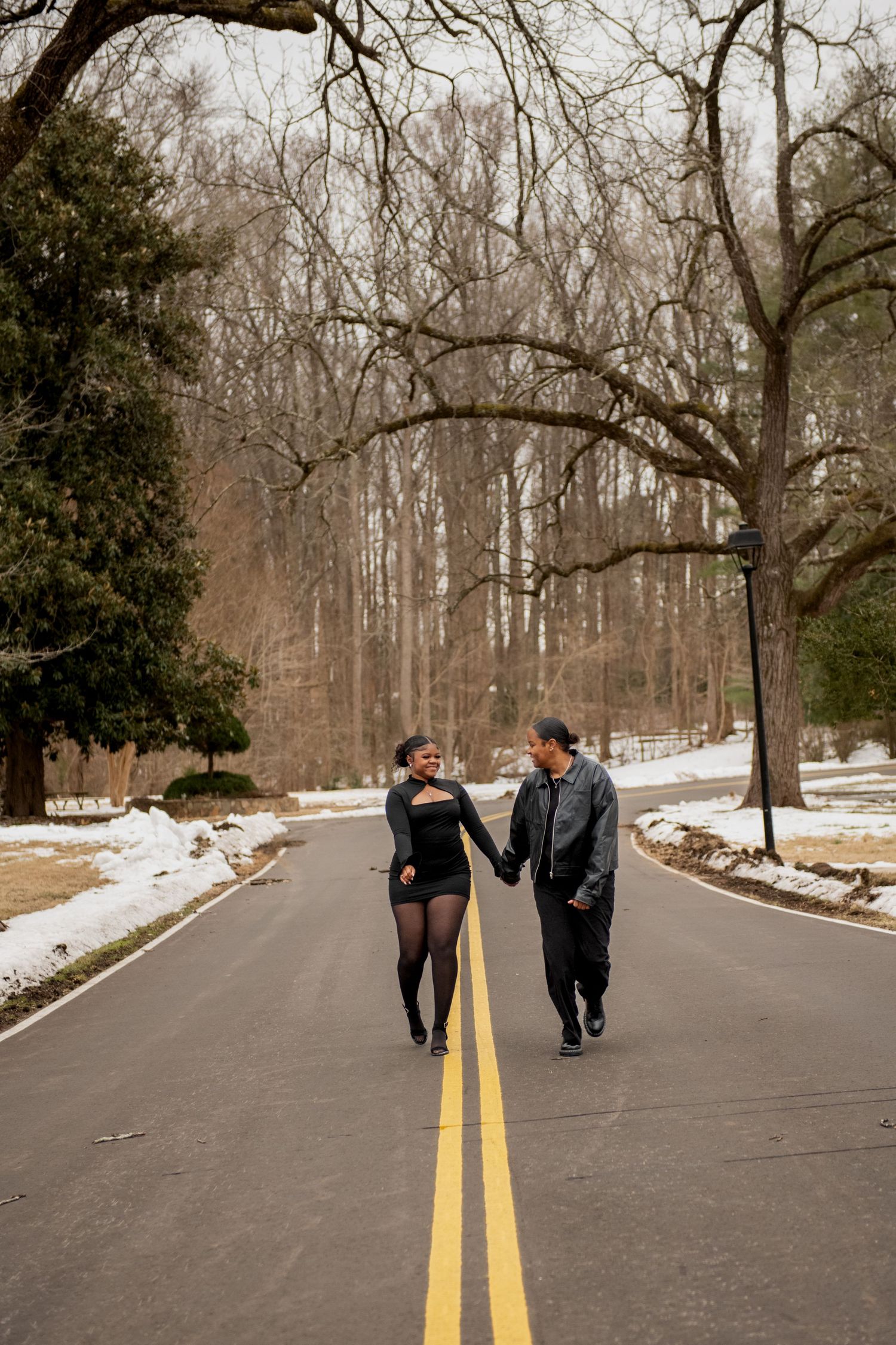 A couple walks hand in hand down the center of a snow-lined road through a winter forest.