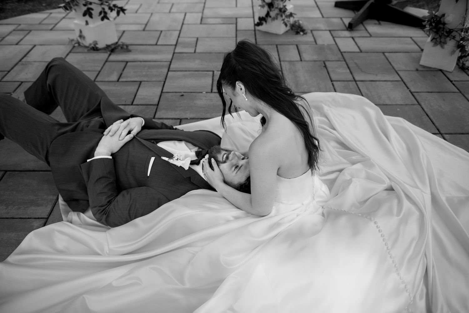 Black and white wedding photo of bride in white gown and groom in suit lying on stone patio, sharing an intimate moment.