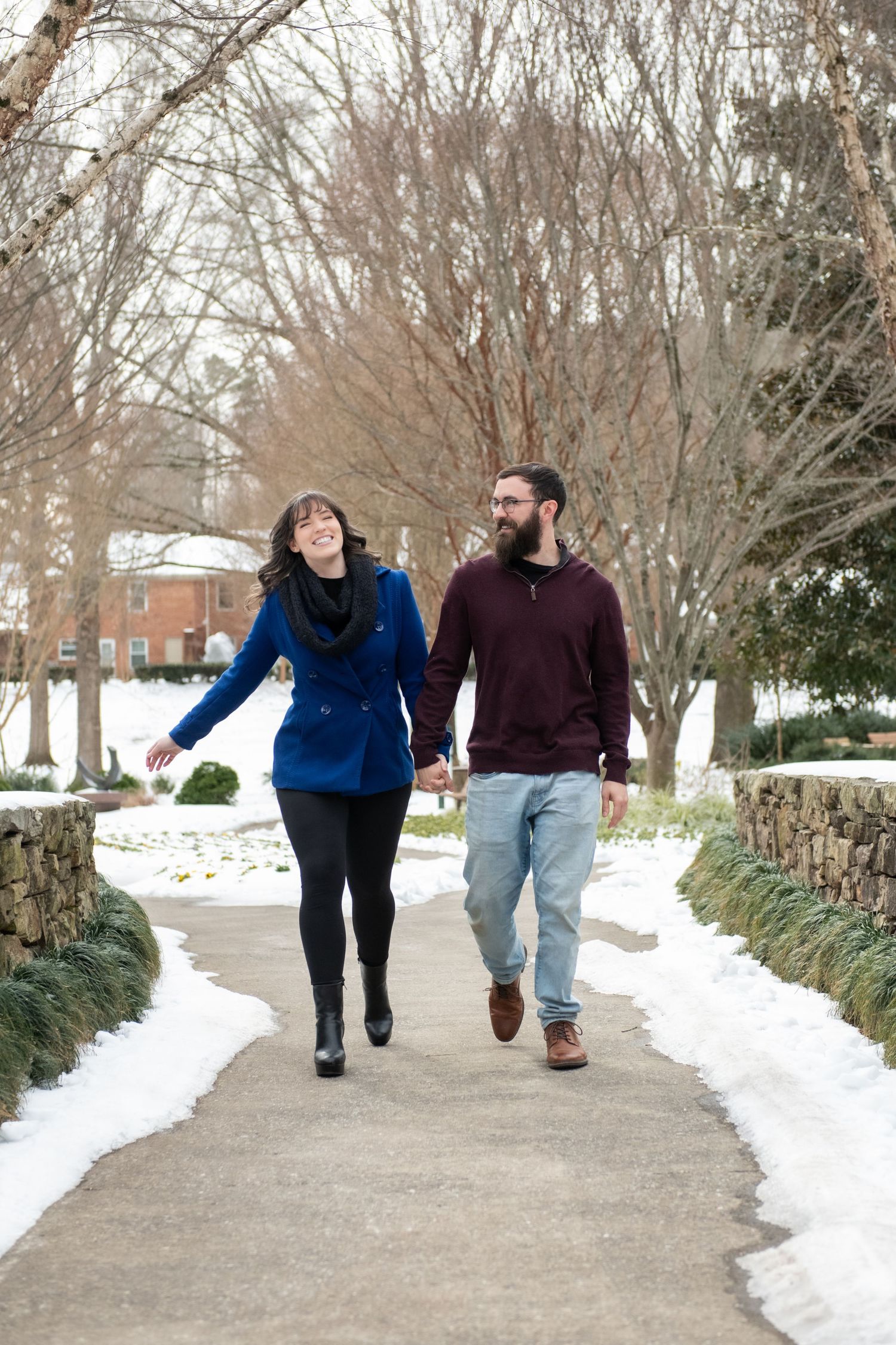 A couple walks together holding hands along a snow-covered pathway surrounded by winter trees.