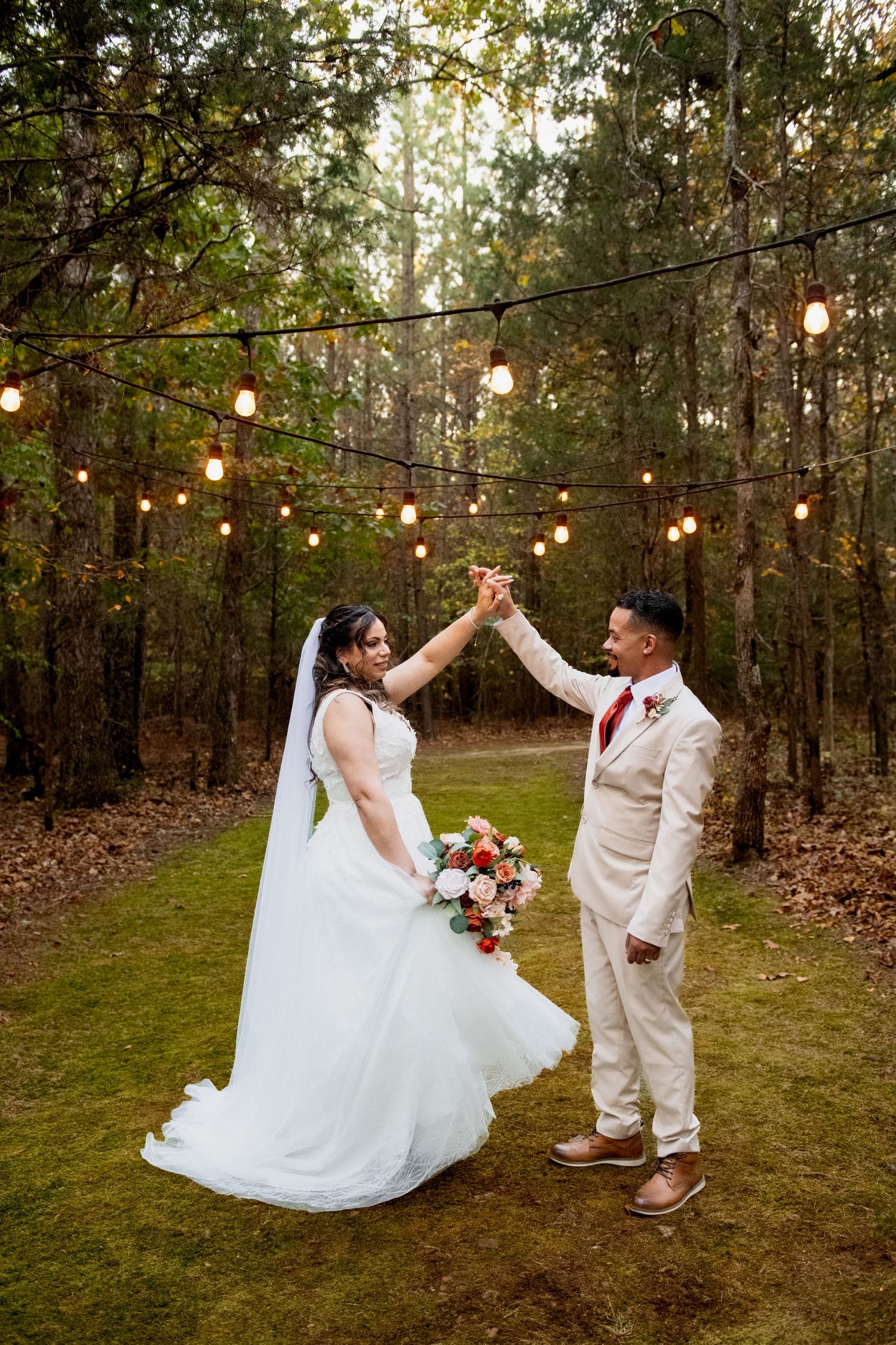 A romantic wedding couple celebrates under string lights in a wooded outdoor setting with a beautiful floral bouquet.