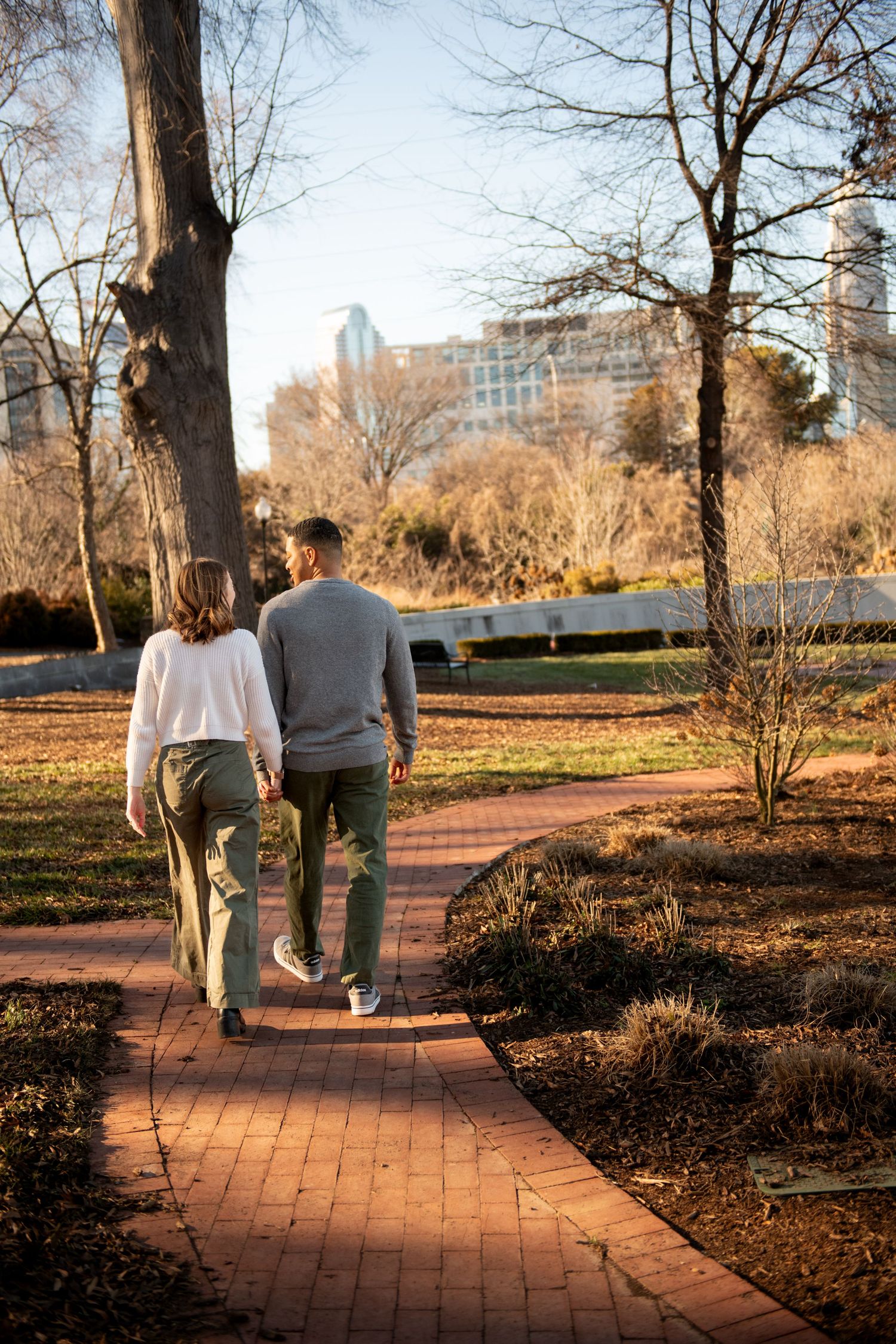 A couple walks along a brick path through a park on a sunny autumn afternoon with city buildings in the background.