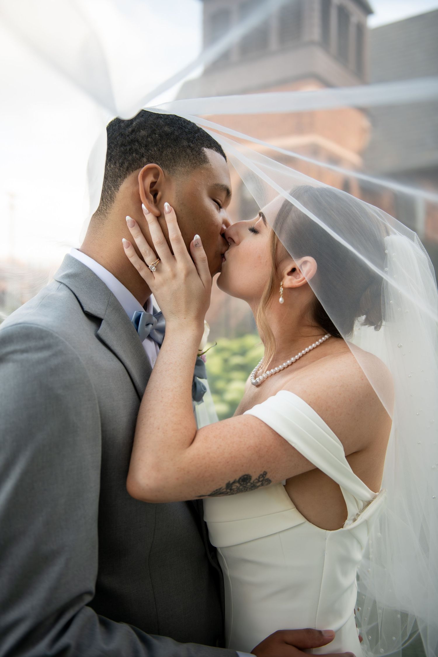 Bride and groom share a romantic kiss under a flowing veil, bride in white dress with pearl necklace, groom in gray suit.