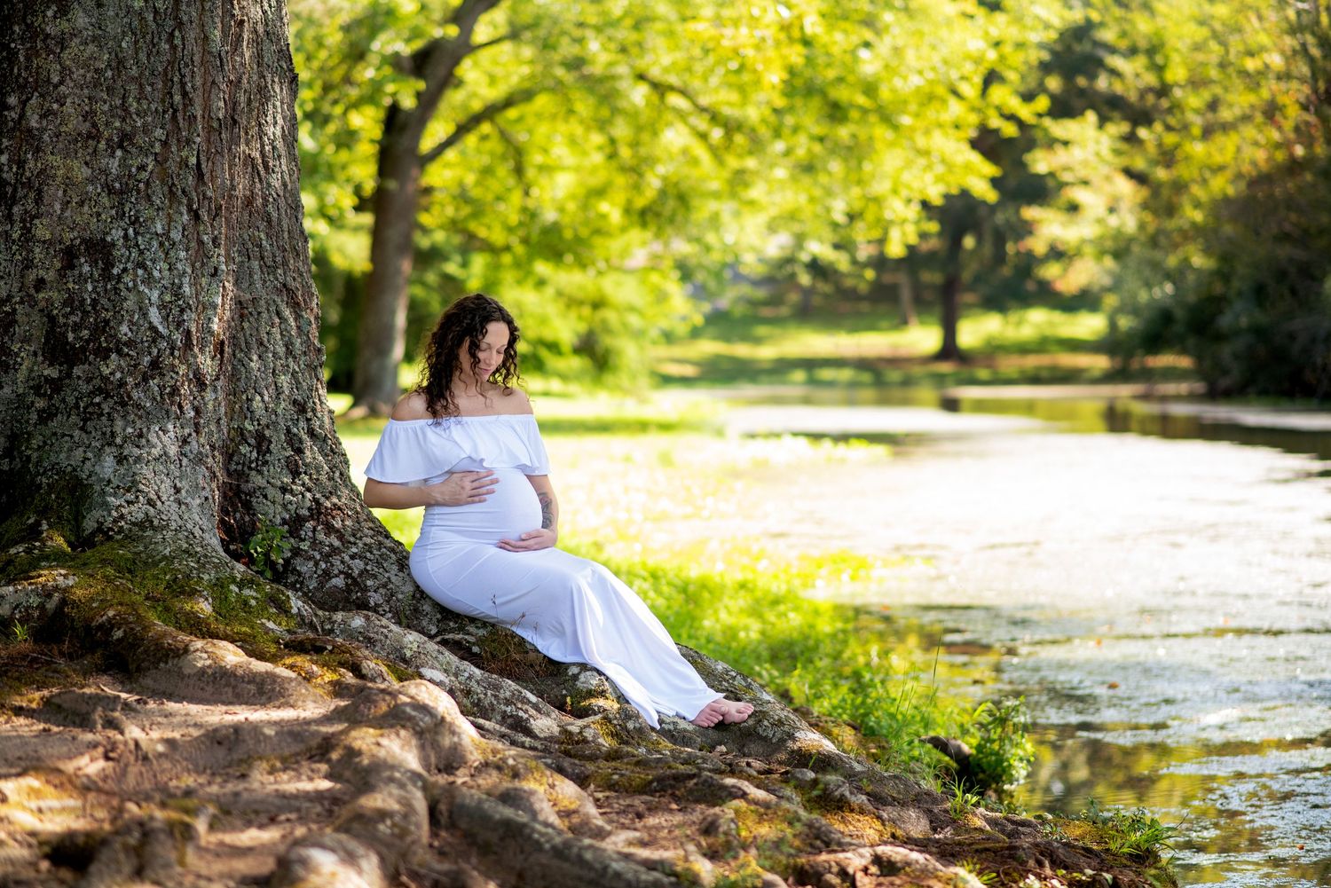 Pregnant woman in white dress sitting by a tree near a serene river in a lush green park on a sunny day.