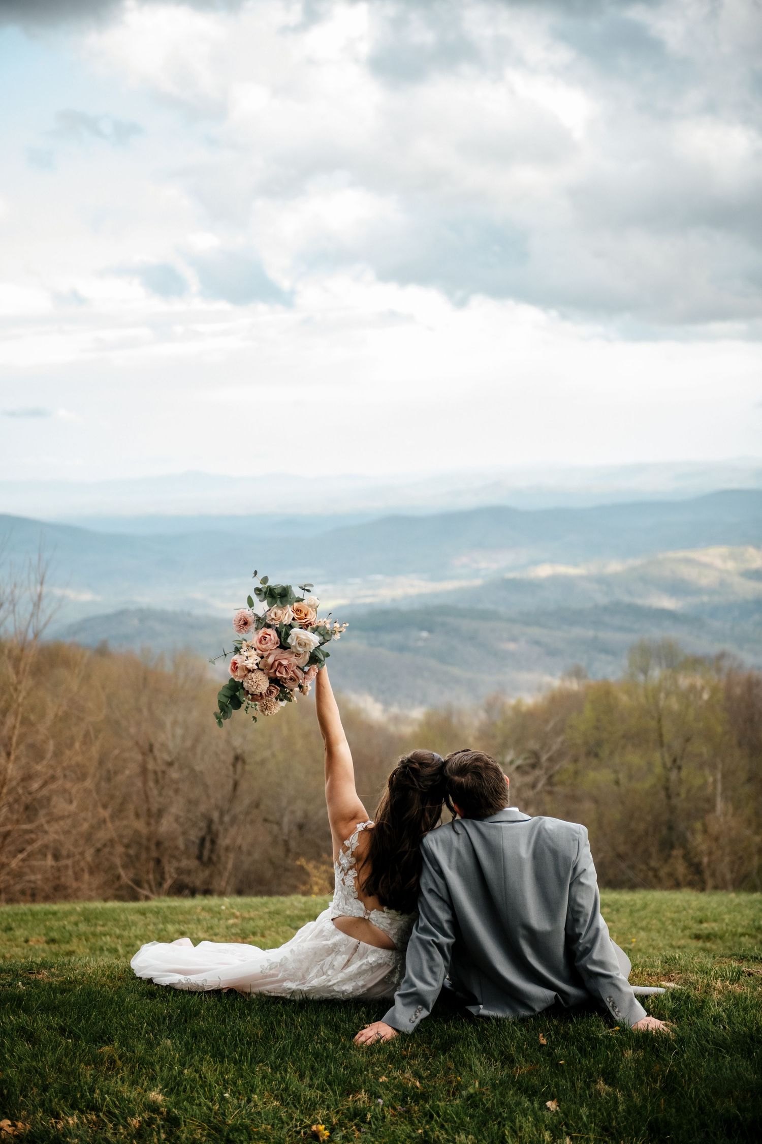 Bride and groom sit on green hilltop, mountains behind them, as bride raises floral bouquet in celebration.