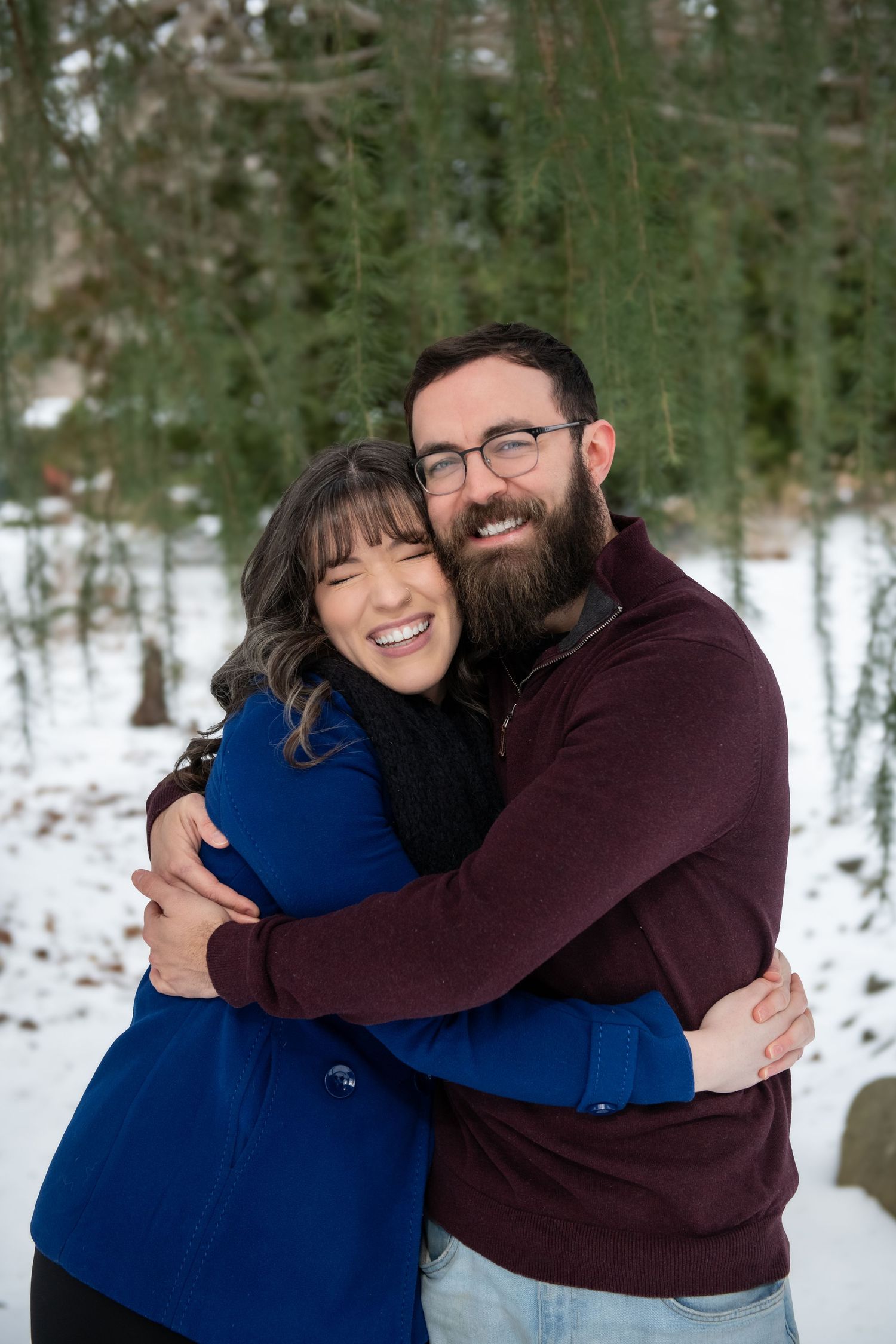 A couple in coordinating blue and burgundy sweaters embrace while smiling in a snowy forest setting.