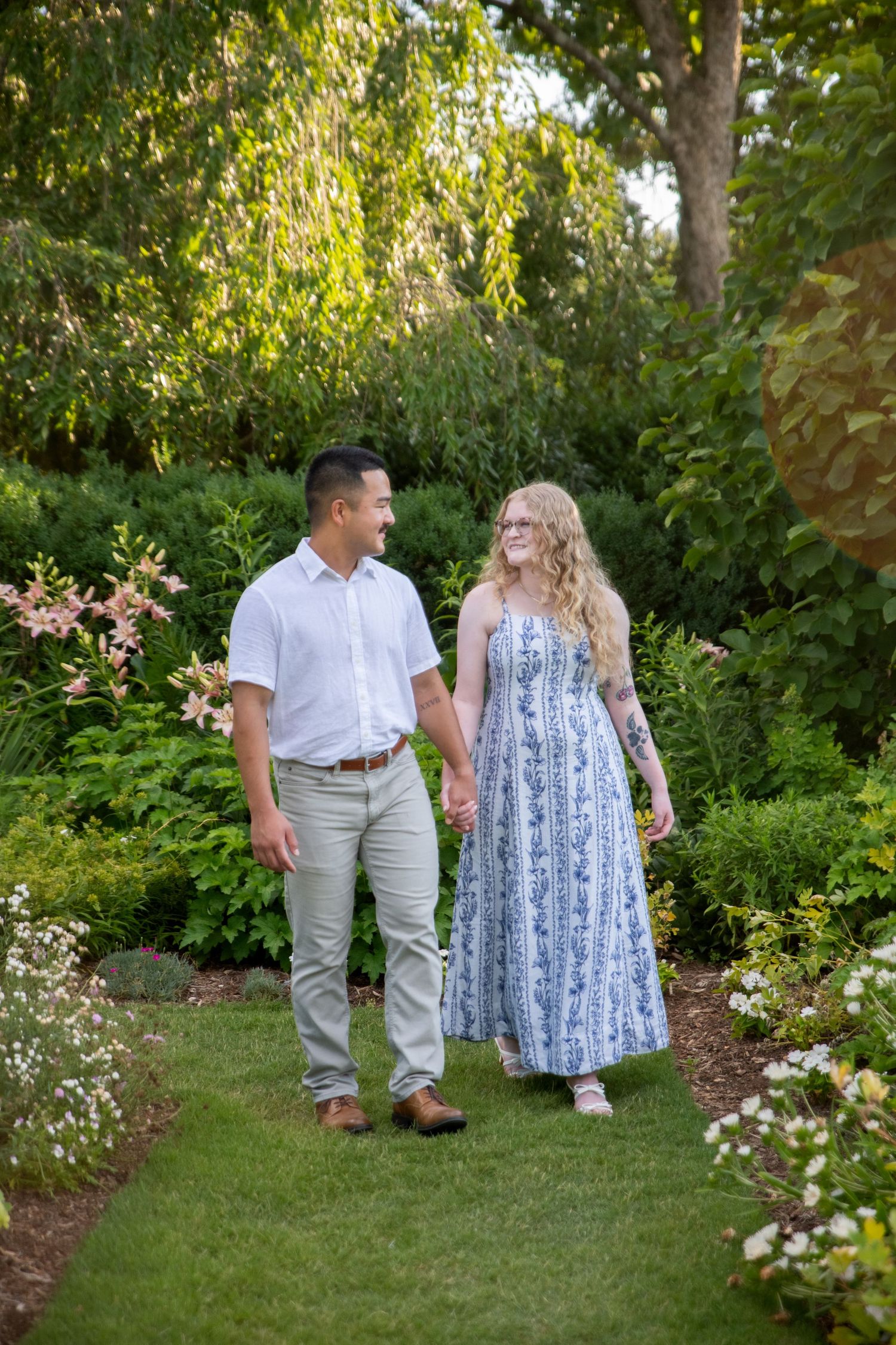 A couple holds hands and smiles while walking through a lush garden path, wearing casual summer attire.