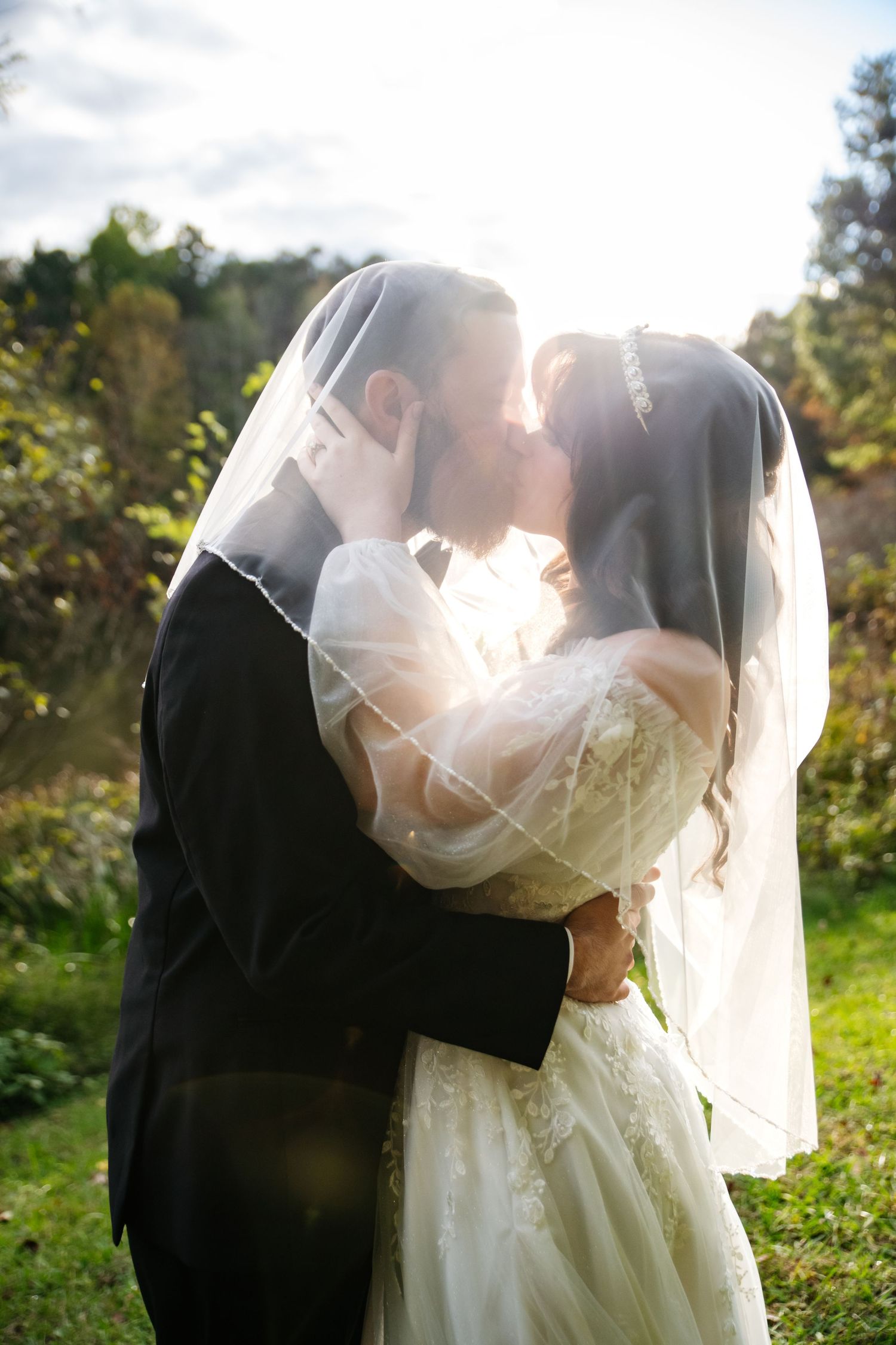 Bride and groom share a romantic kiss under a sheer wedding veil in a sunlit outdoor garden with autumn trees behind them.