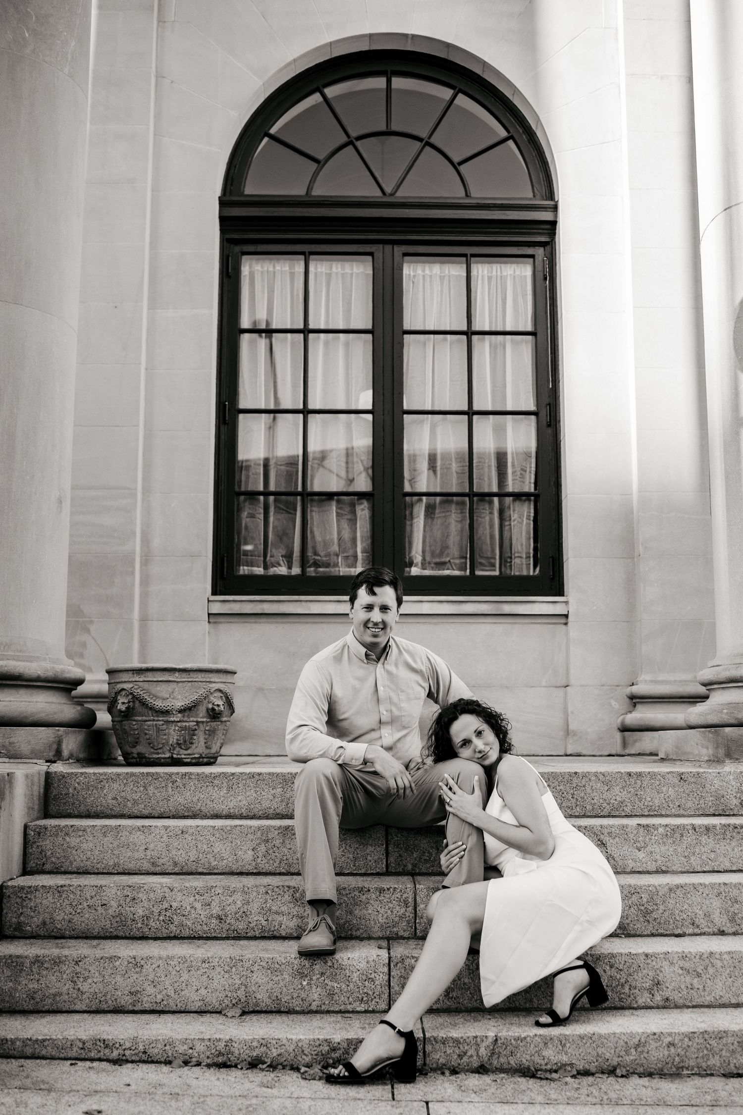 A couple sitting on stone steps beneath an arched window of a classical building.