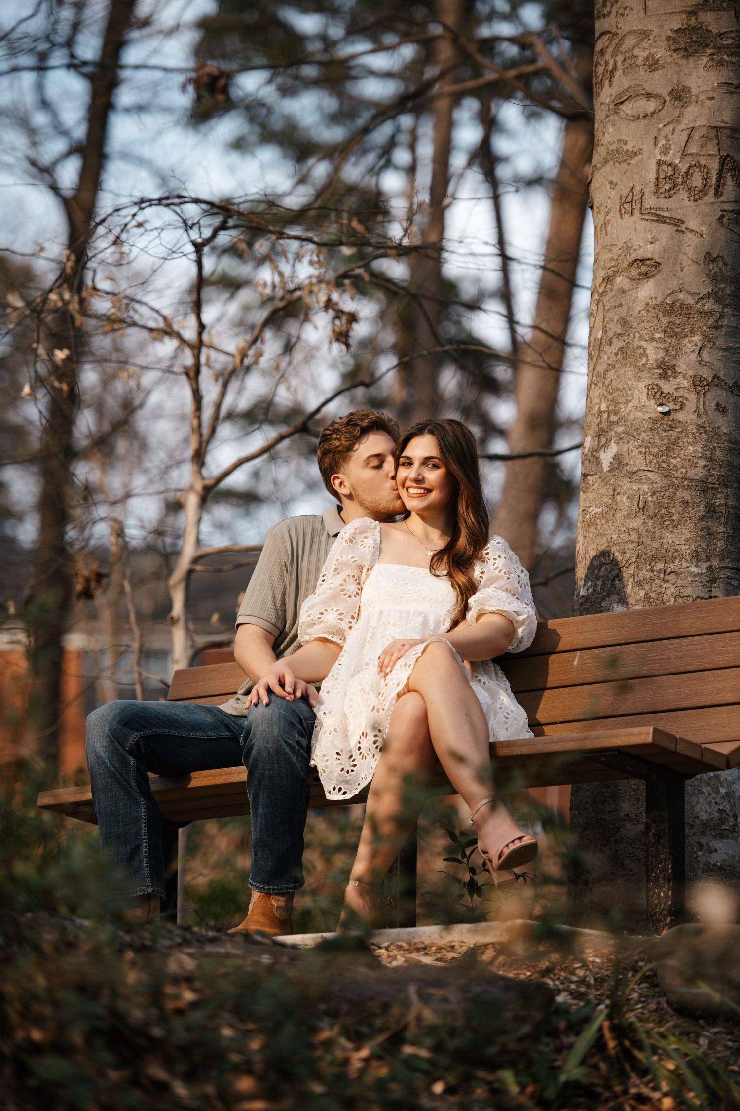 A romantic couple cuddles on a wooden bench in a sunlit forest setting.