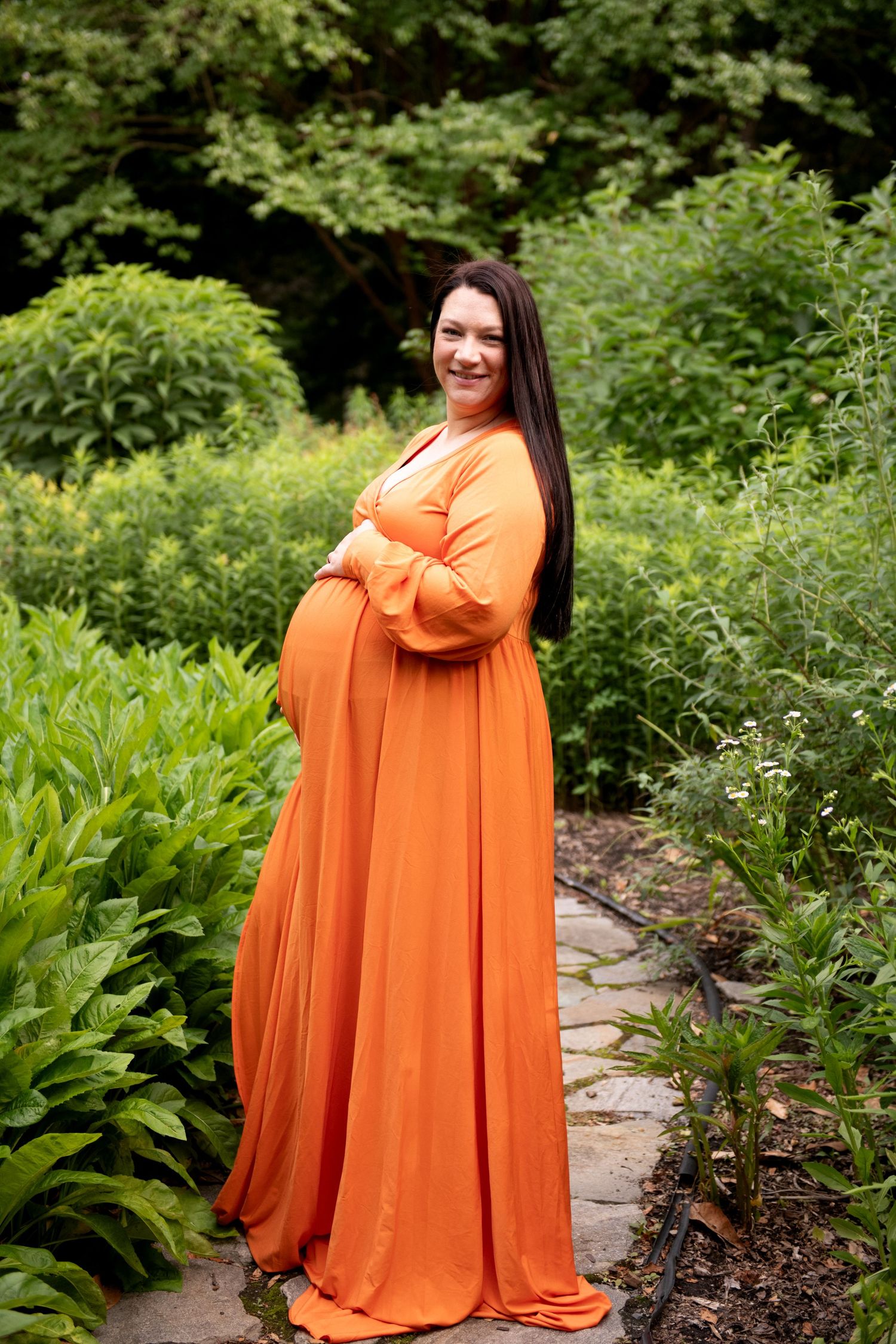 Smiling pregnant woman in orange maternity gown posing outdoors in a lush green garden.