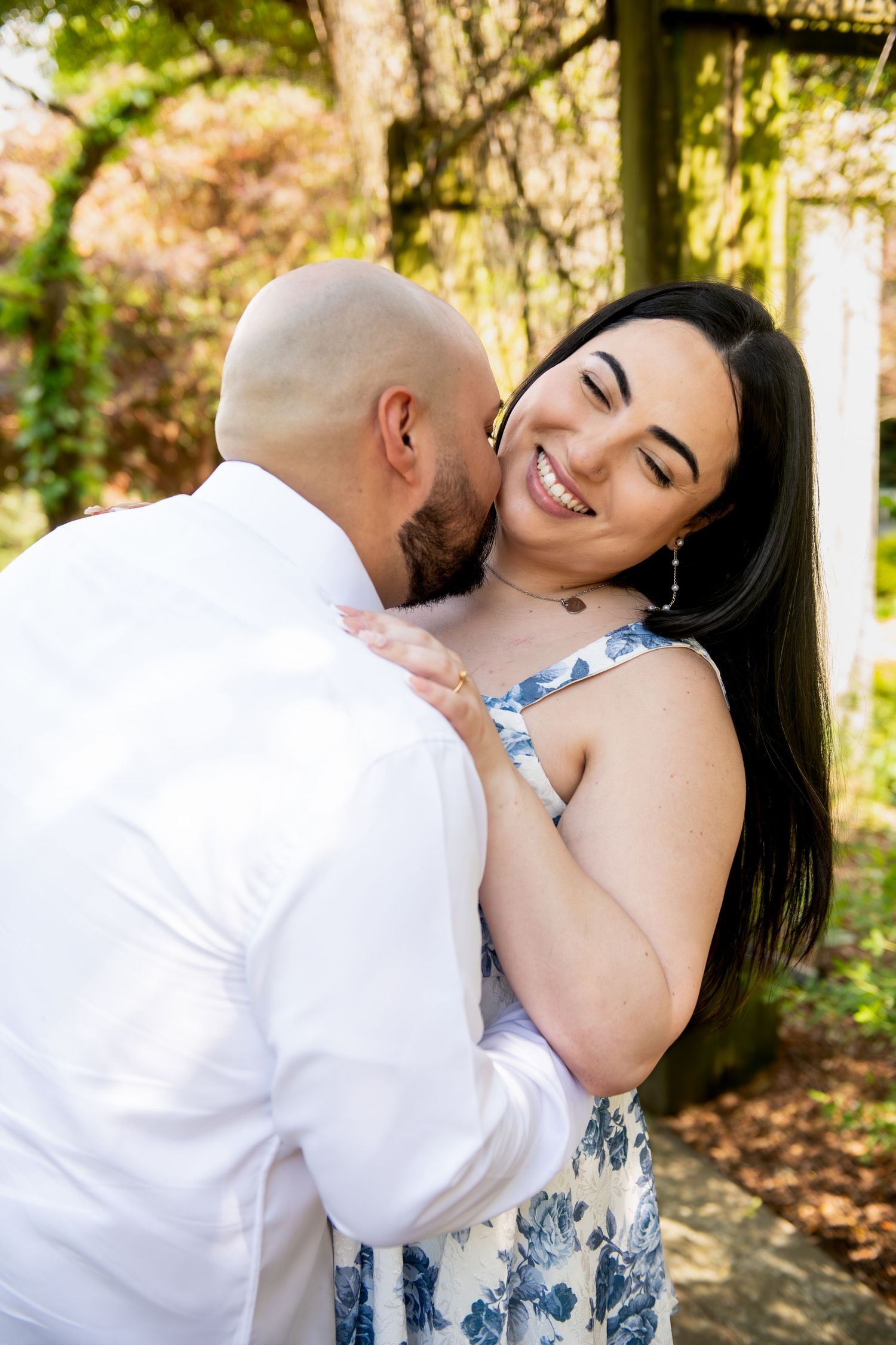 A smiling woman in a floral dress embraces a bald man in a white shirt outdoors among autumn trees.