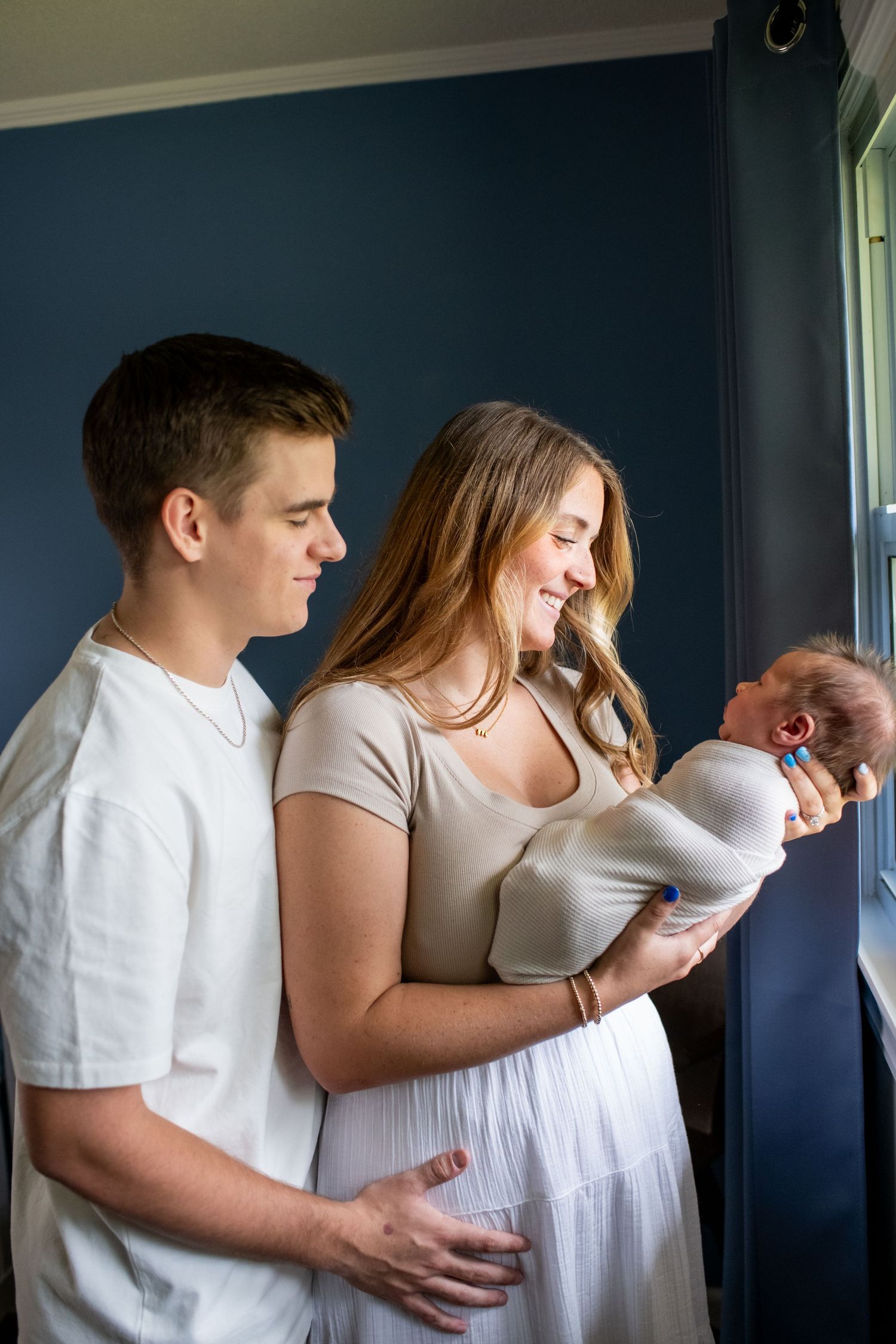 A tender moment captured in natural light by a window as a couple embraces their newborn baby.