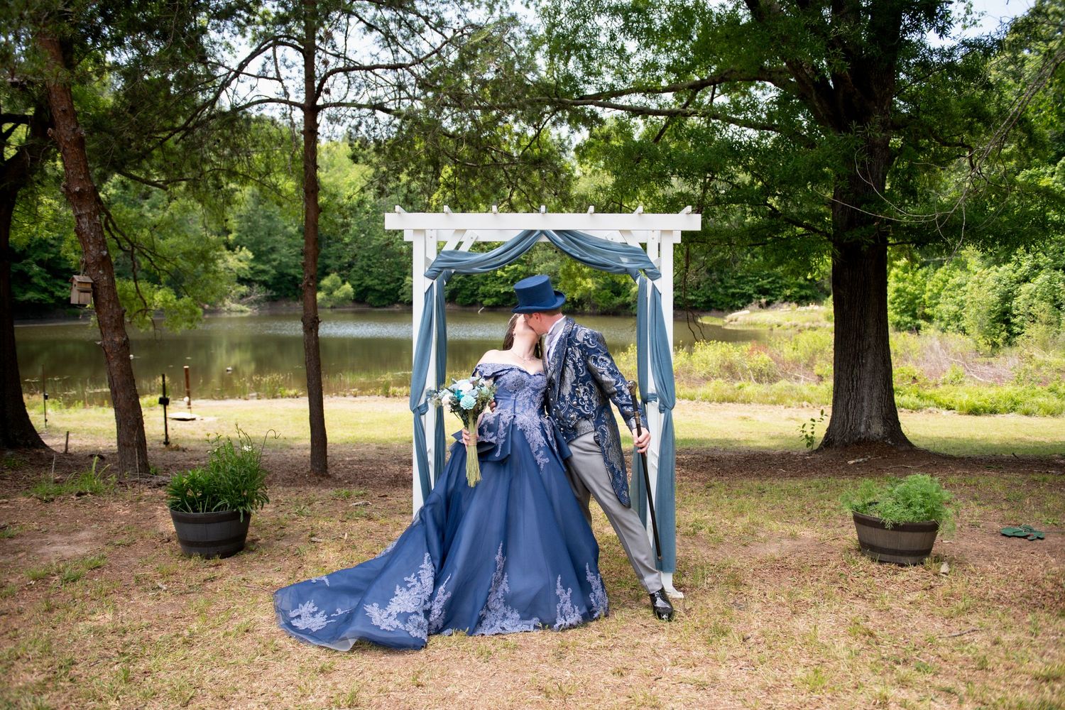 Woman in blue gown stands at outdoor wedding arch by a lake, surrounded by lush green trees.