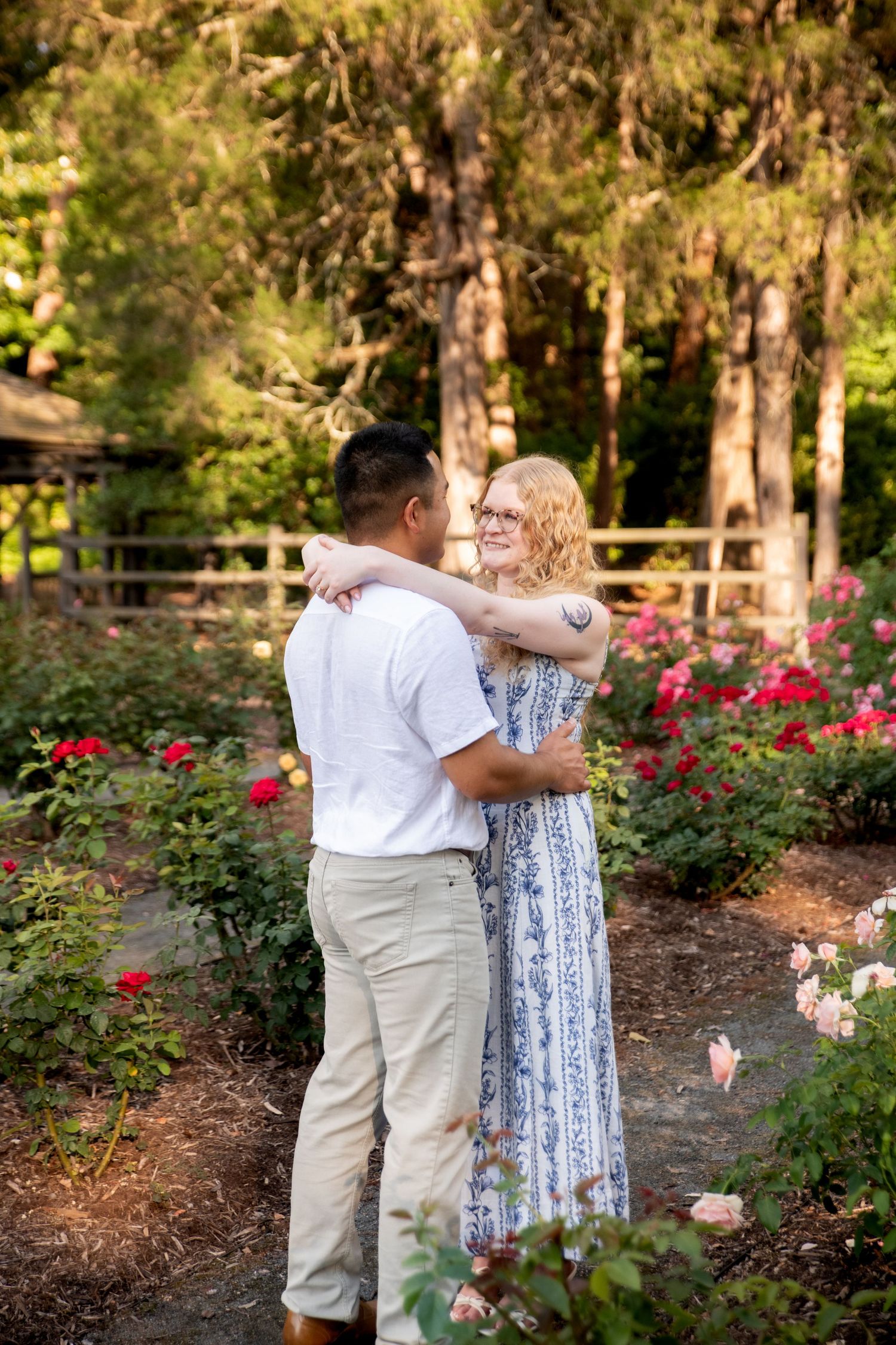 Couple embracing in a rose garden, woman in blue floral dress smiling at man in white shirt amid colorful blooms.