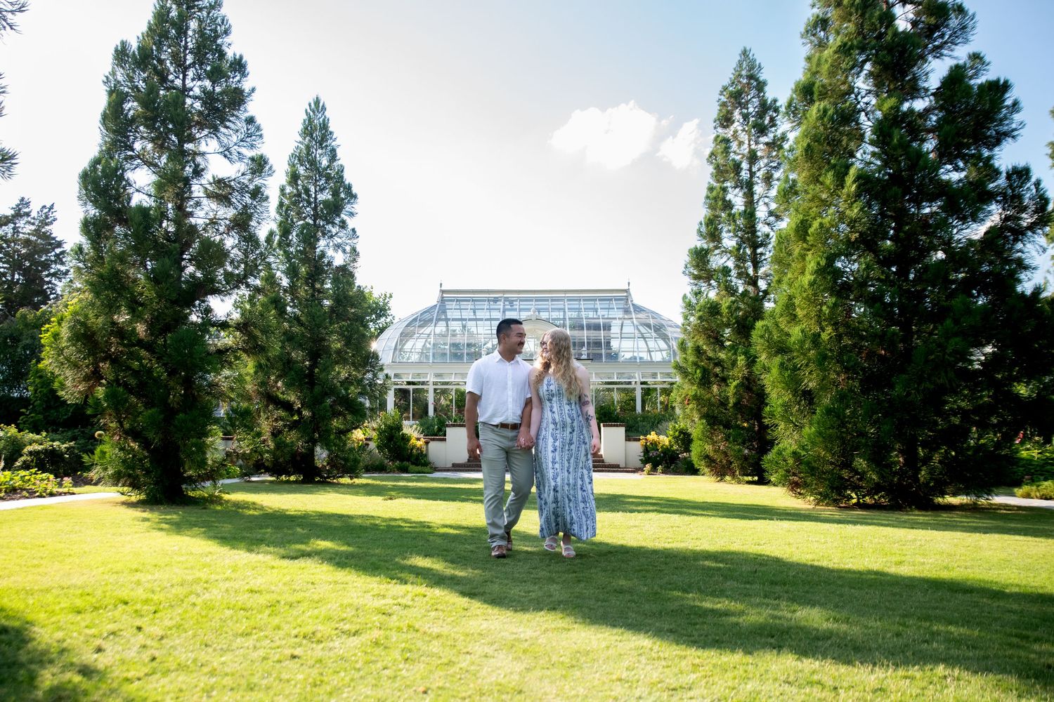 Couple standing on green lawn in front of a glass greenhouse surrounded by tall evergreen trees on a sunny day.