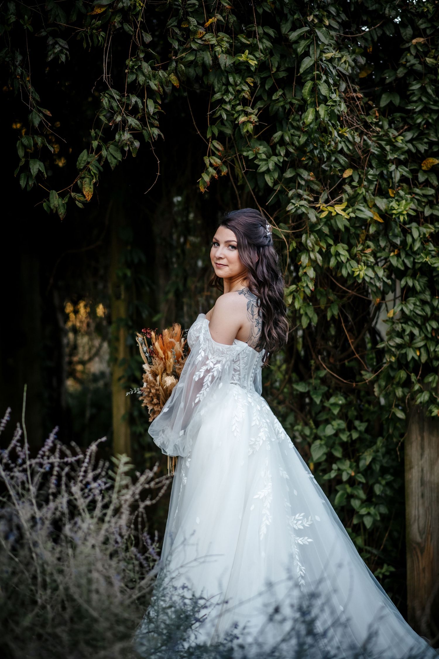 Smiling bride in white lace off-shoulder gown holding dried bouquet, posing in lush green garden archway.