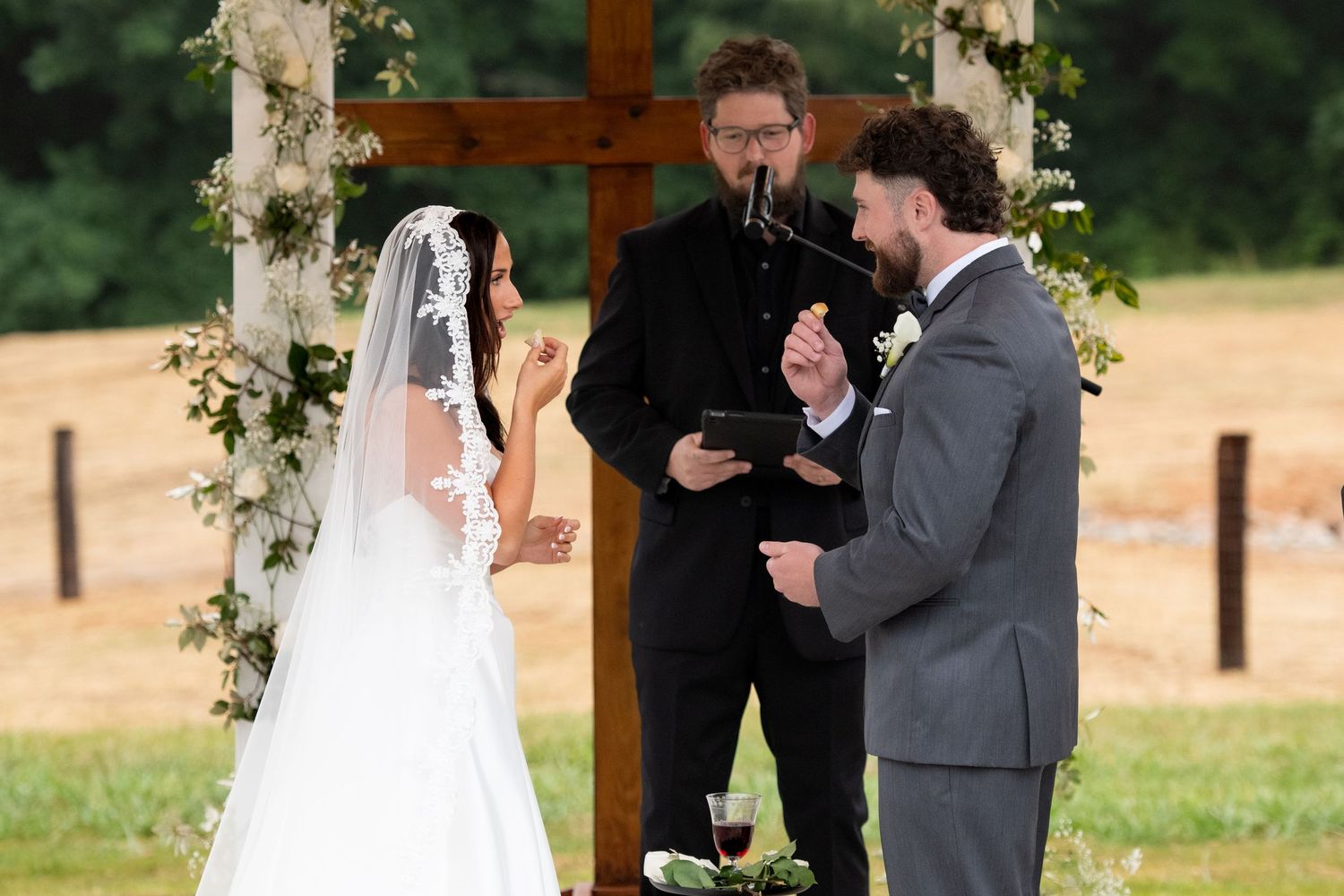 Outdoor wedding ceremony with bride in white veil, groom in gray suit, and officiant holding tablet before wooden cross.