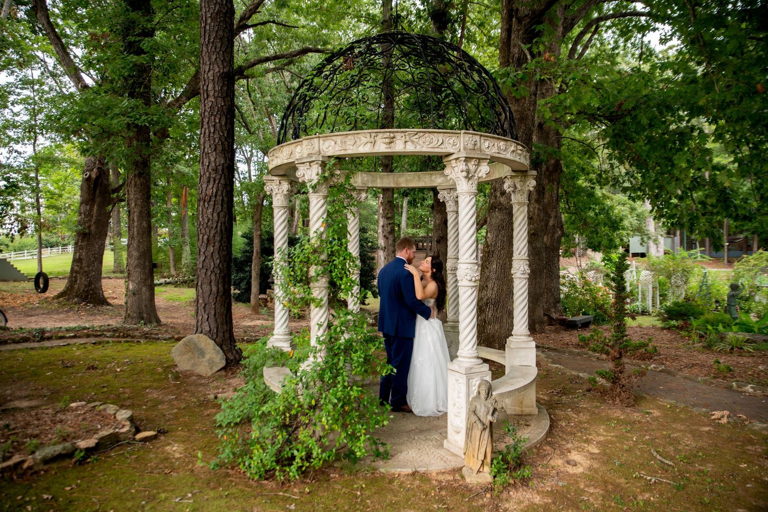 Bride in a white gown and groom in a dark suit embrace beneath an ornate white gazebo with columns, surrounded by lush green trees.