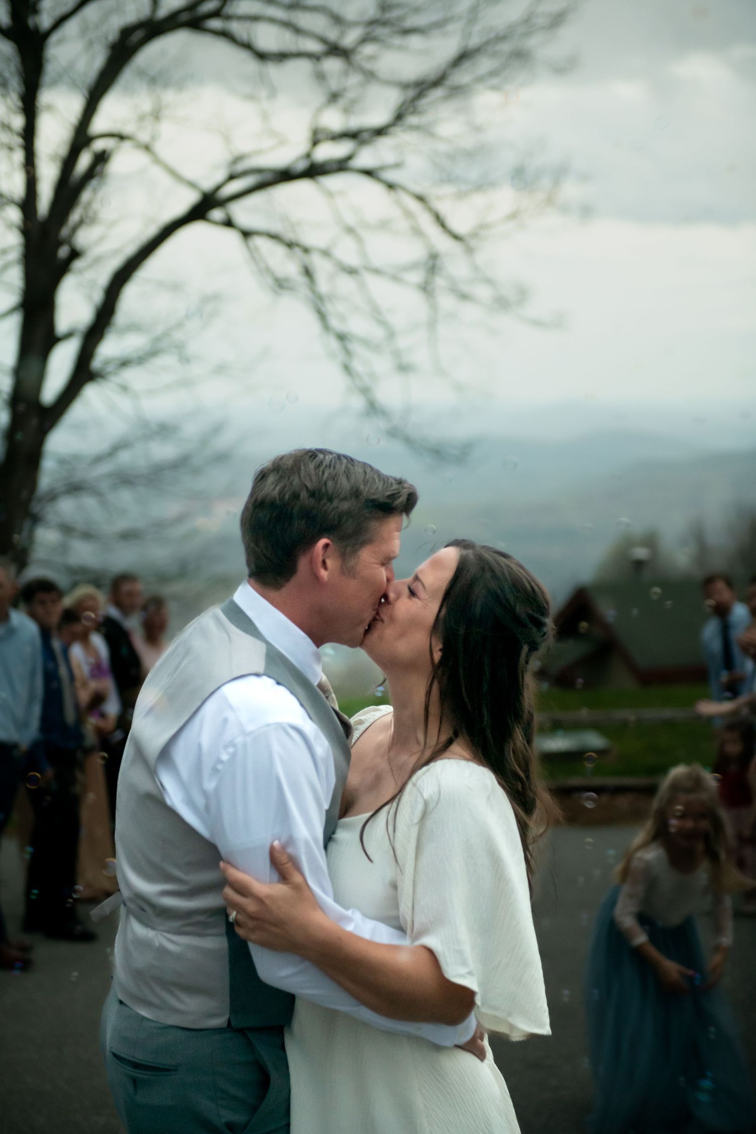 A bride and groom share their first kiss outdoors at a rustic wedding with guests celebrating in the background.