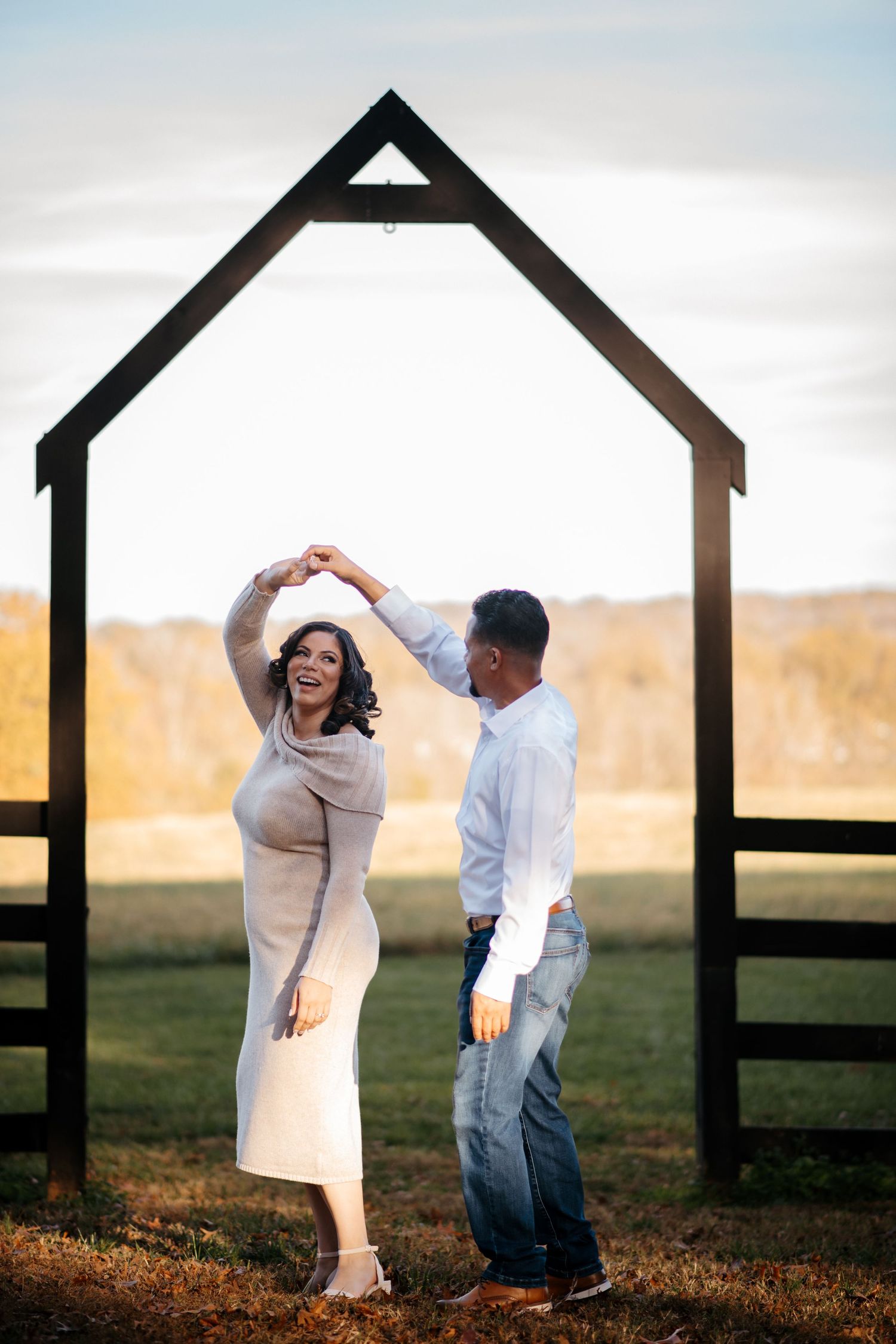 Couple dancing and twirling beneath house-shaped frames in a field at sunset.