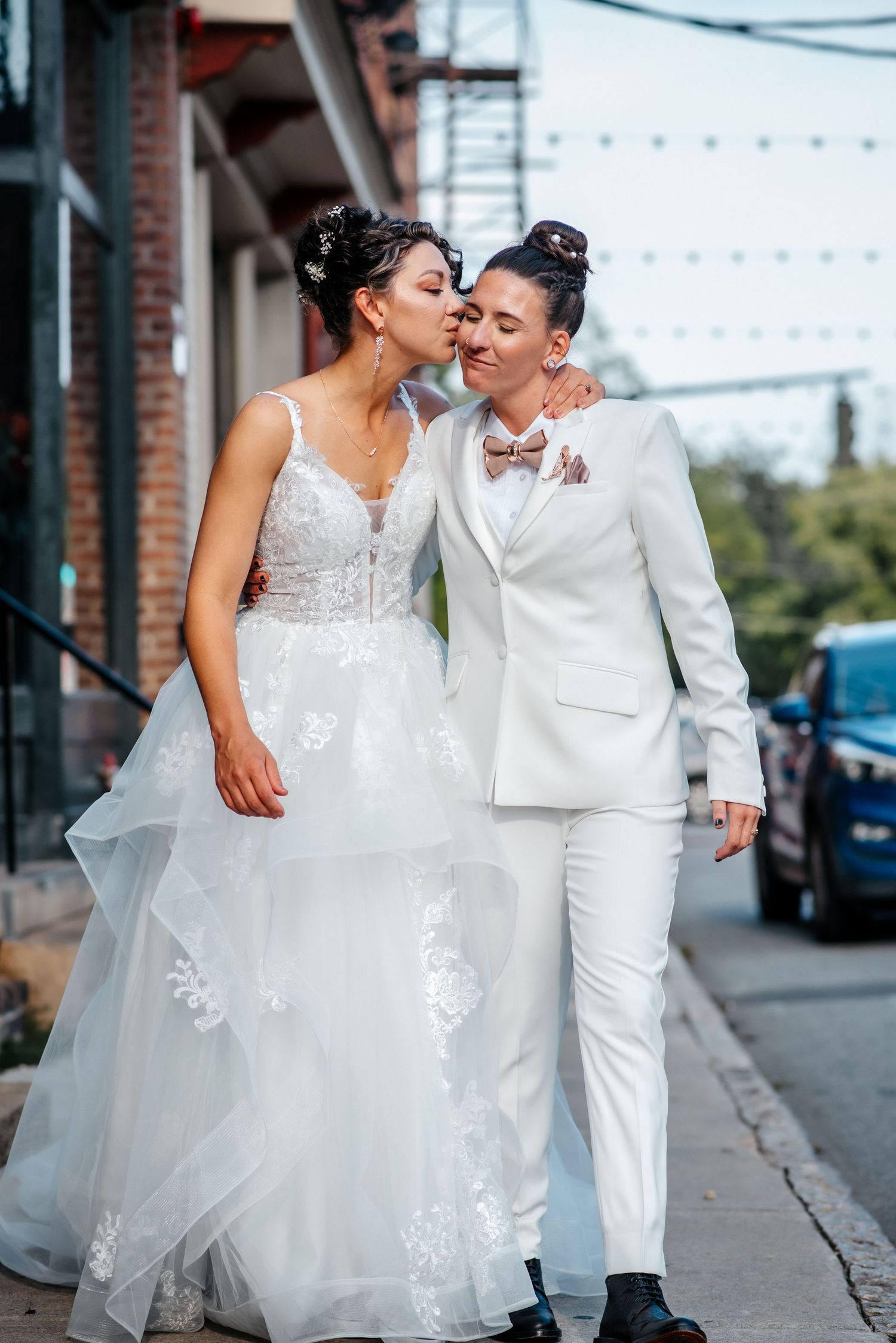 Two brides in white wedding attire share a kiss on a city sidewalk, one in a gown and one in a suit.