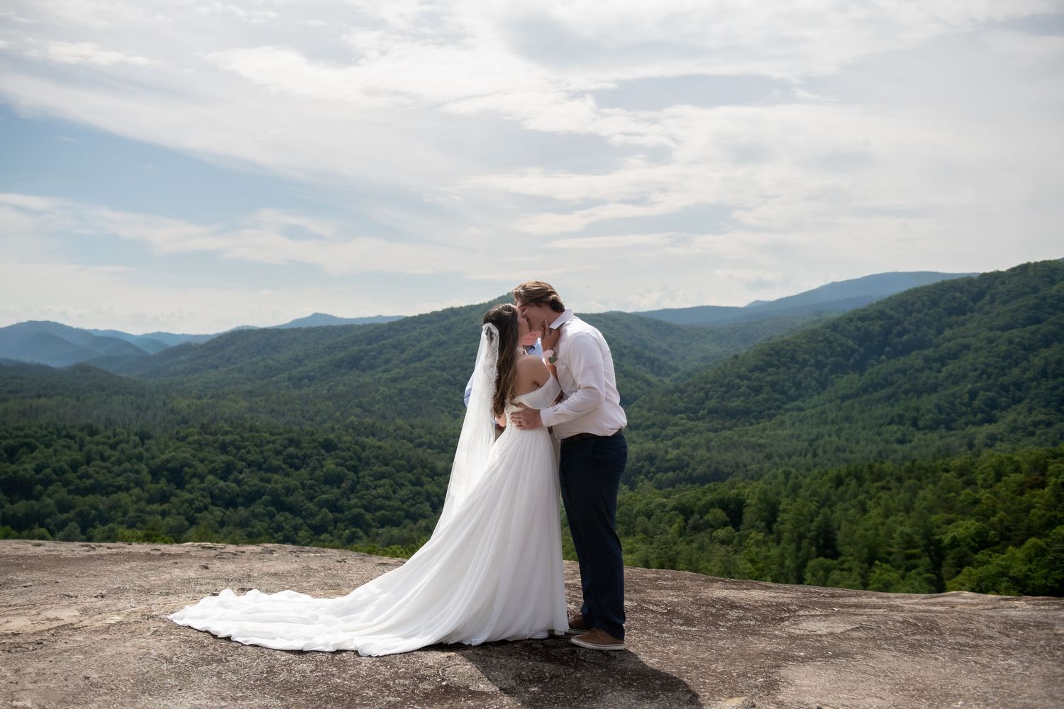 Bride and groom kissing on a rocky mountain overlook with lush green hills and cloudy sky backdrop.