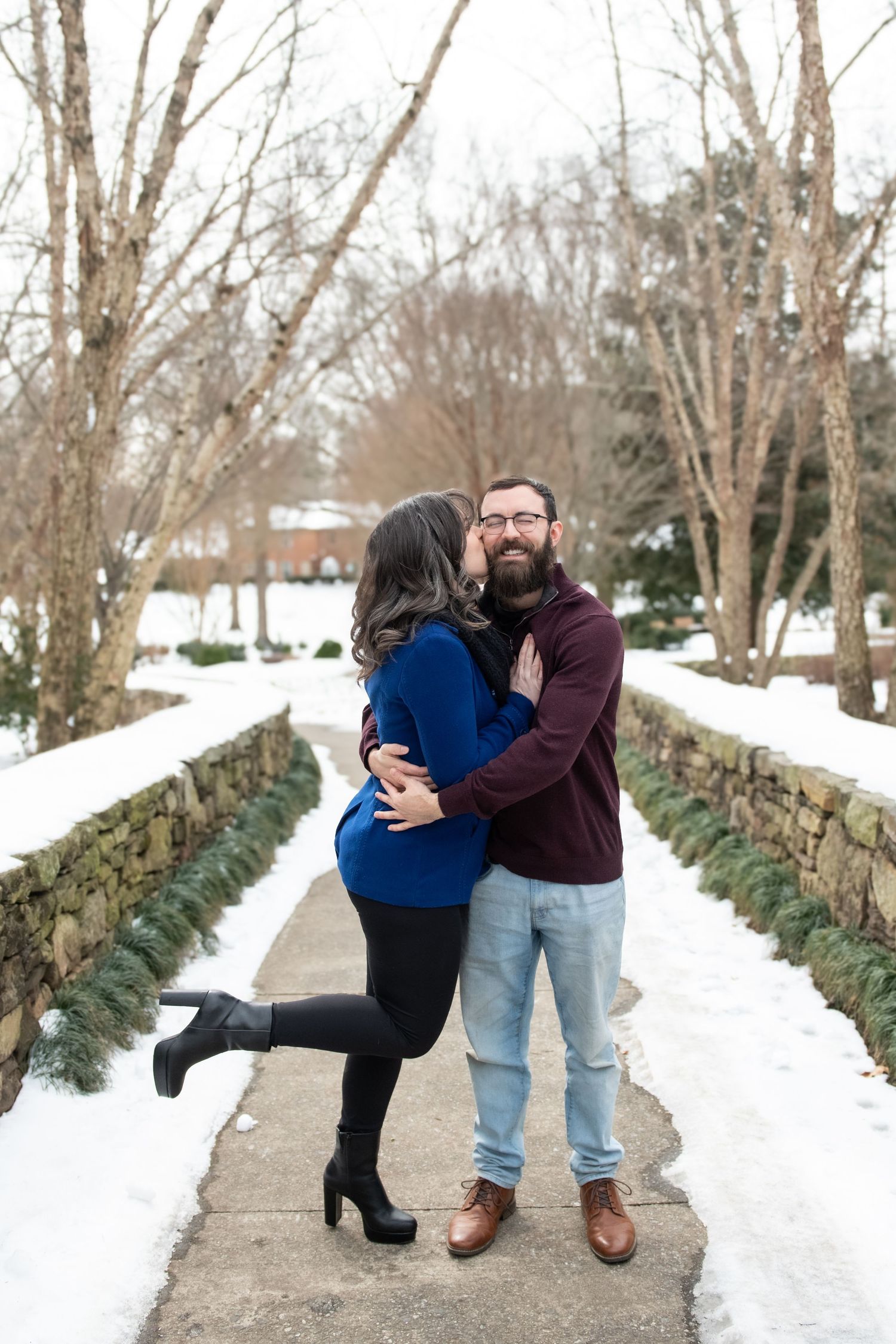 A couple embraces on a snowy winter path lined with evergreen garland.