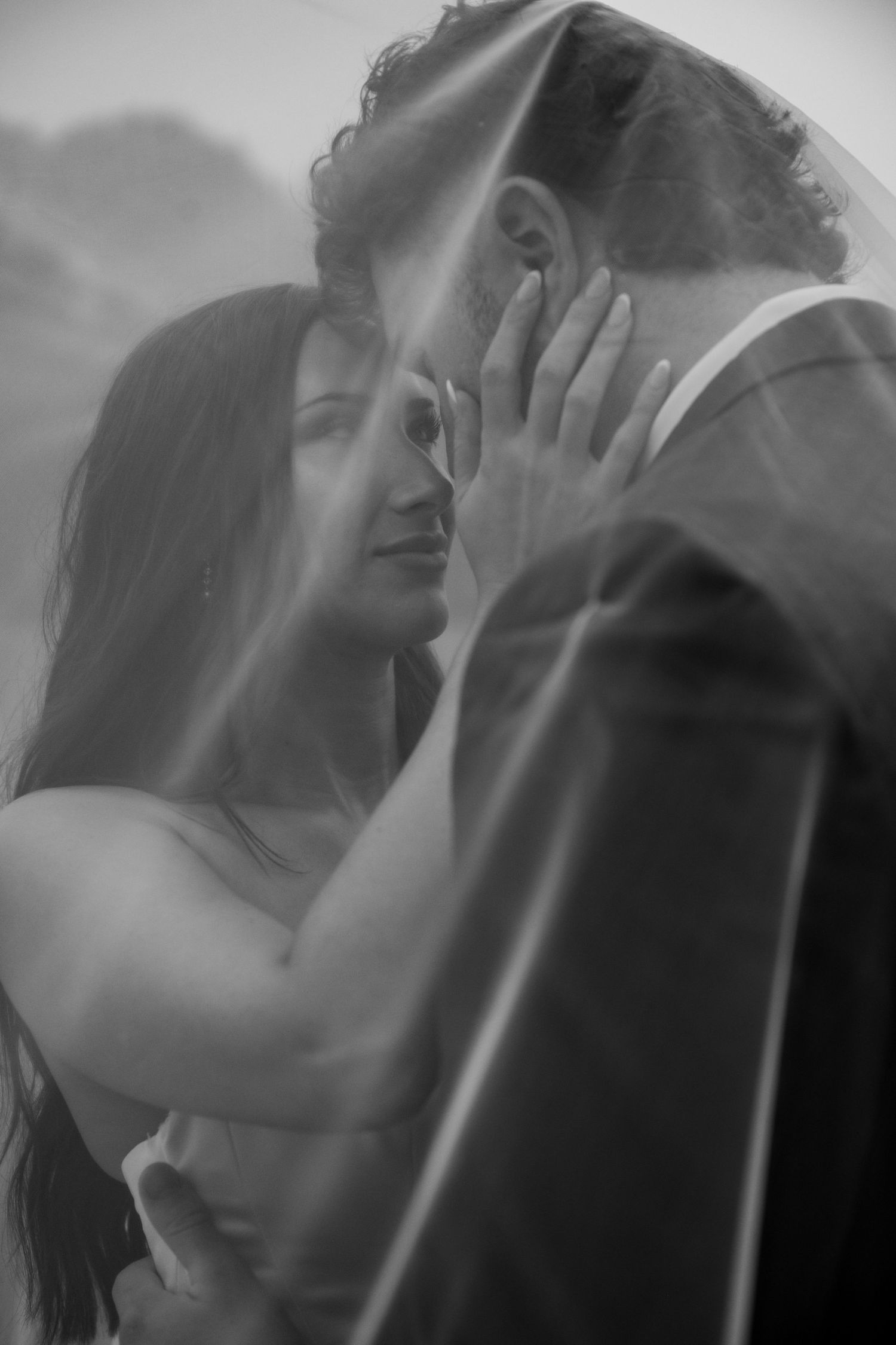 Black and white photo of a couple sharing an intimate moment under a veil, bride touching grooms face tenderly.