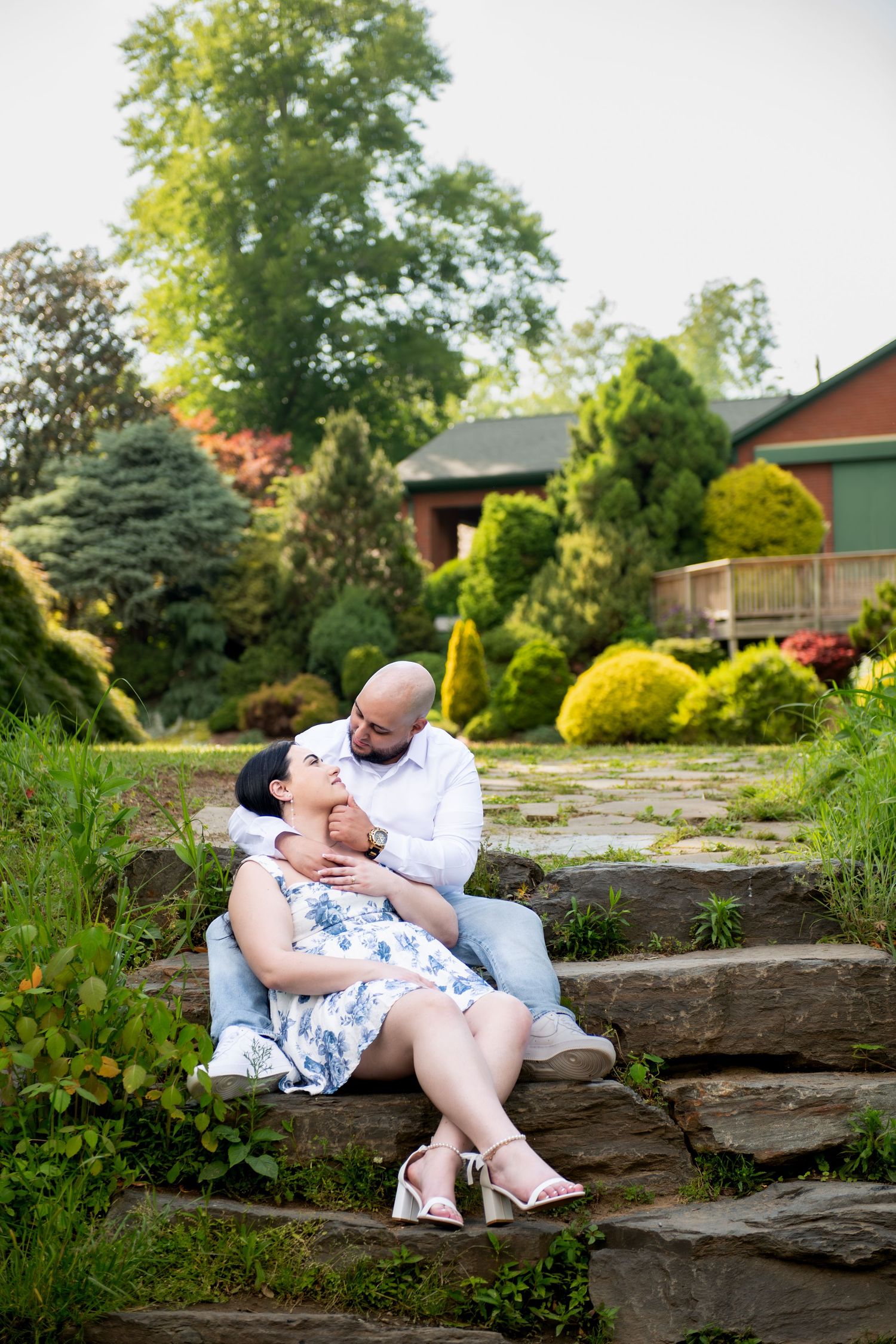 Couple sitting on stone steps in a lush garden, woman in floral dress leaning back against man in white shirt.