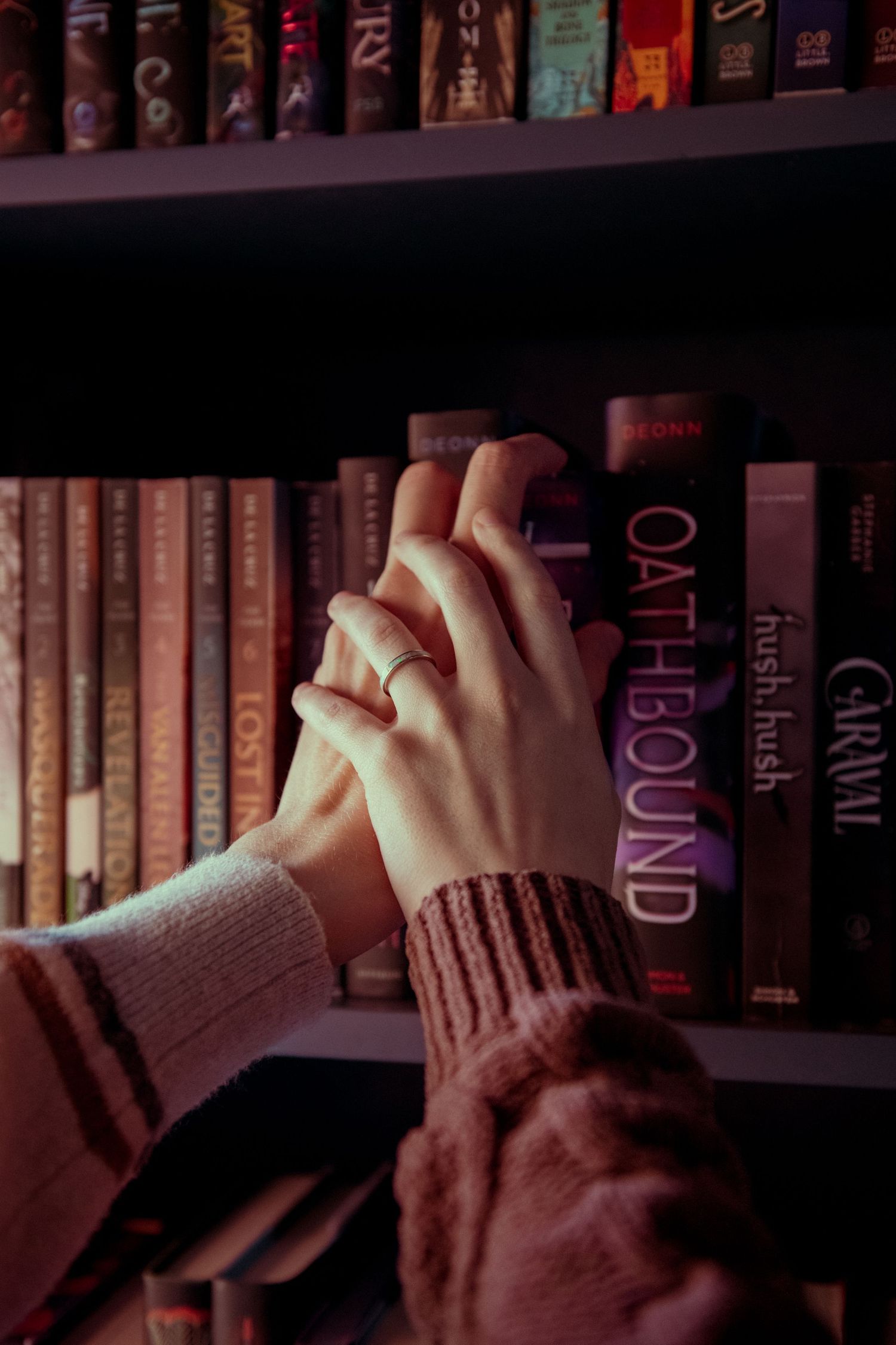 Two hands intertwined near a bookshelf featuring titles like Oathbound and Hush Hush, with one hand wearing a silver ring.
