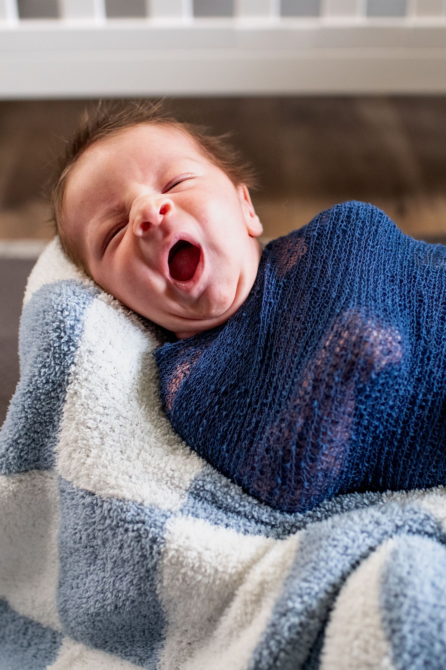 A baby wrapped in blue and gray blankets yawns while lying down.