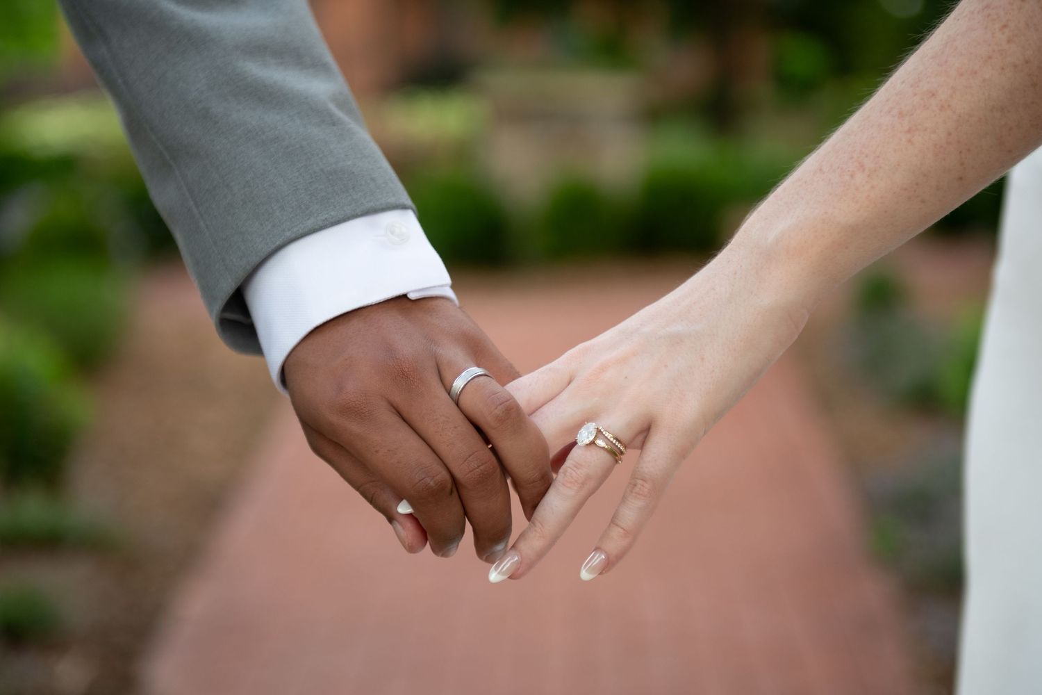 Interracial couple holding hands showing wedding and engagement rings on a brick pathway.