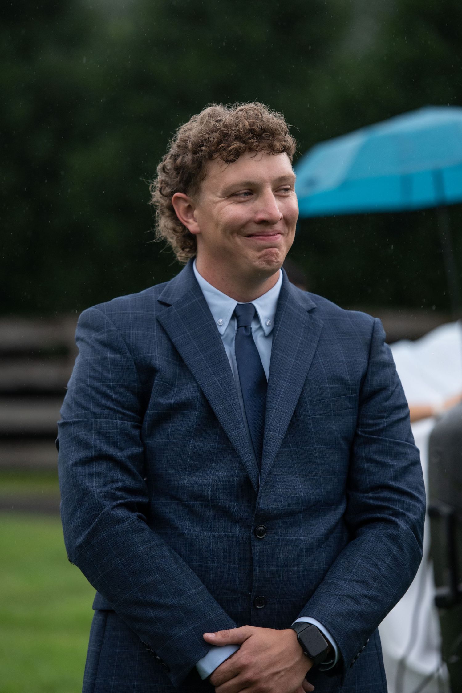 Young man with curly hair smiling in a navy blue suit and tie at an outdoor event on a rainy day.