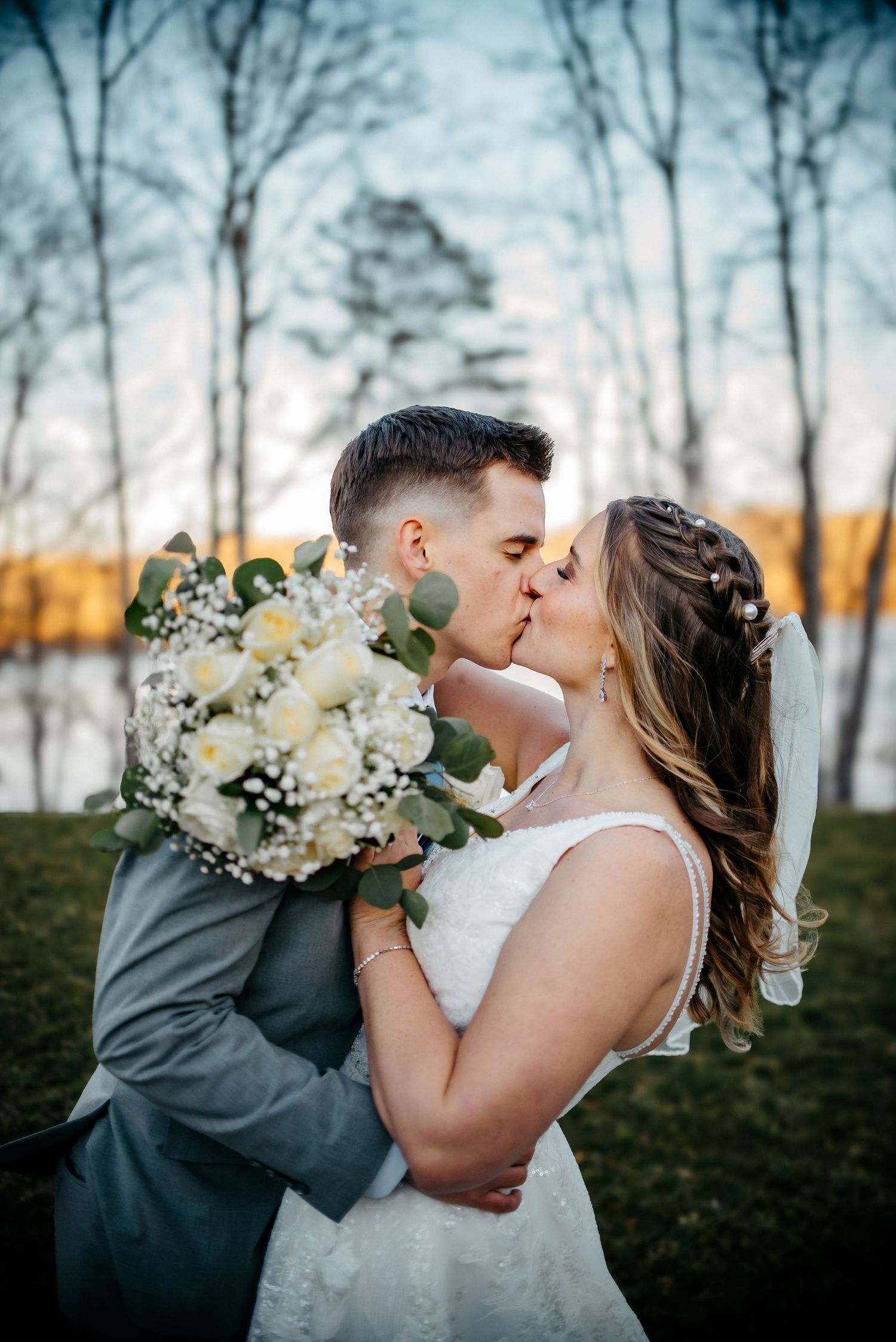 A romantic wedding couple shares tender kisses while holding a white and green floral bouquet at sunset.